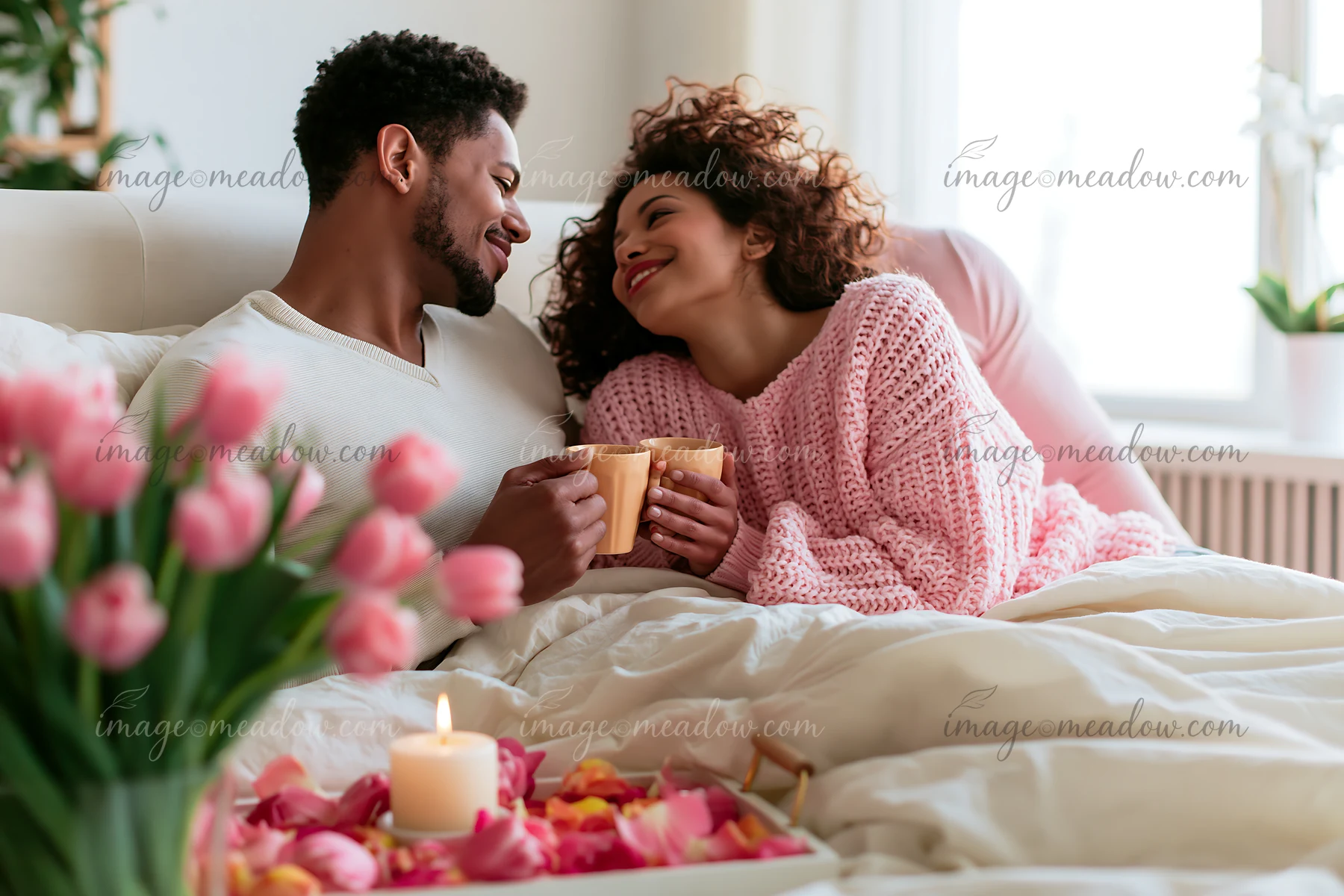 Smiling couple shares coffee in bed beside pink tulips, rose petals, and a candle in a bright bedroom.