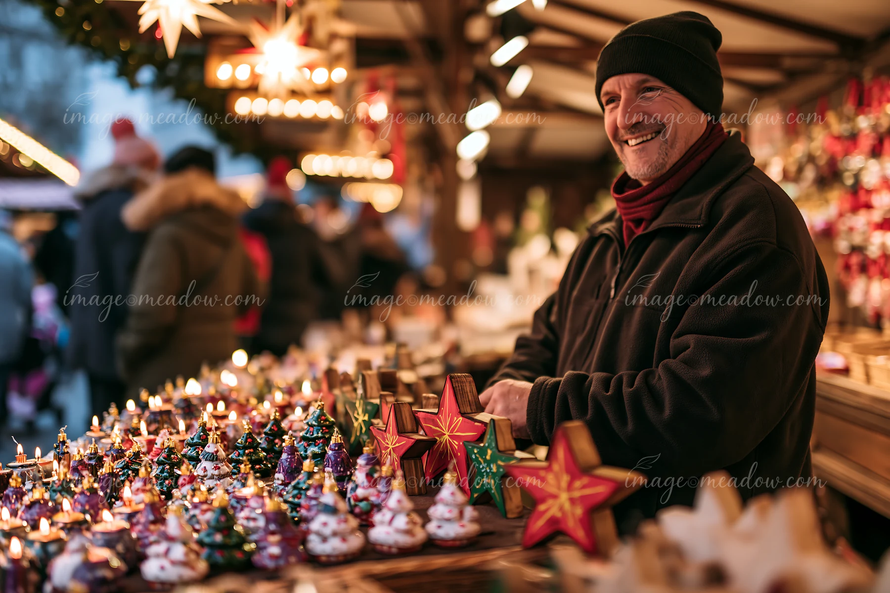 Smiling vendor stands behind a warmly lit Christmas market stall displaying handmade ornaments.