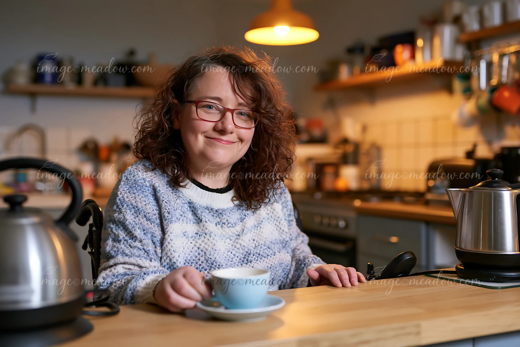 Smiling woman wearing glasses sits at a kitchen counter with coffee in a warmly lit home kitchen.