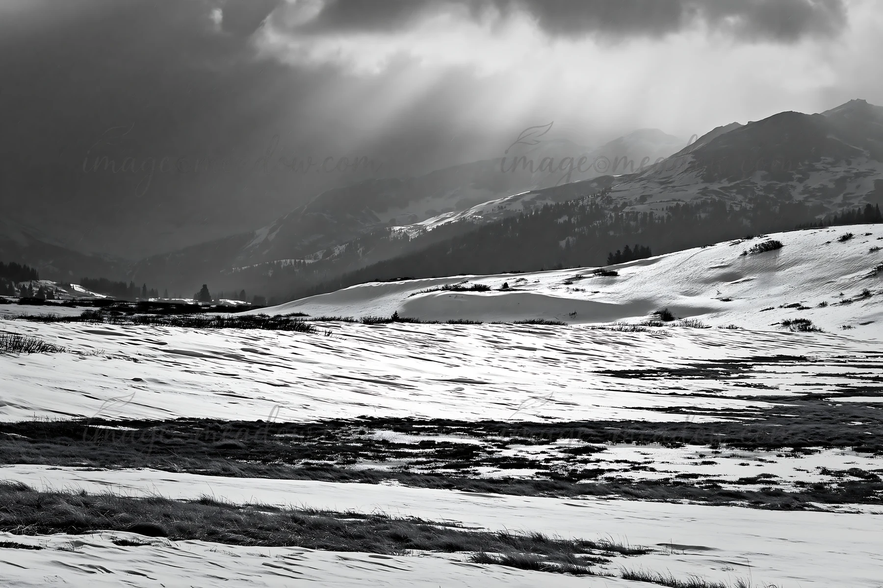 A snow-covered meadow stretches across the landscape with mountains in the background. Dark clouds loom above, casting shadows on the white ground, creating a u