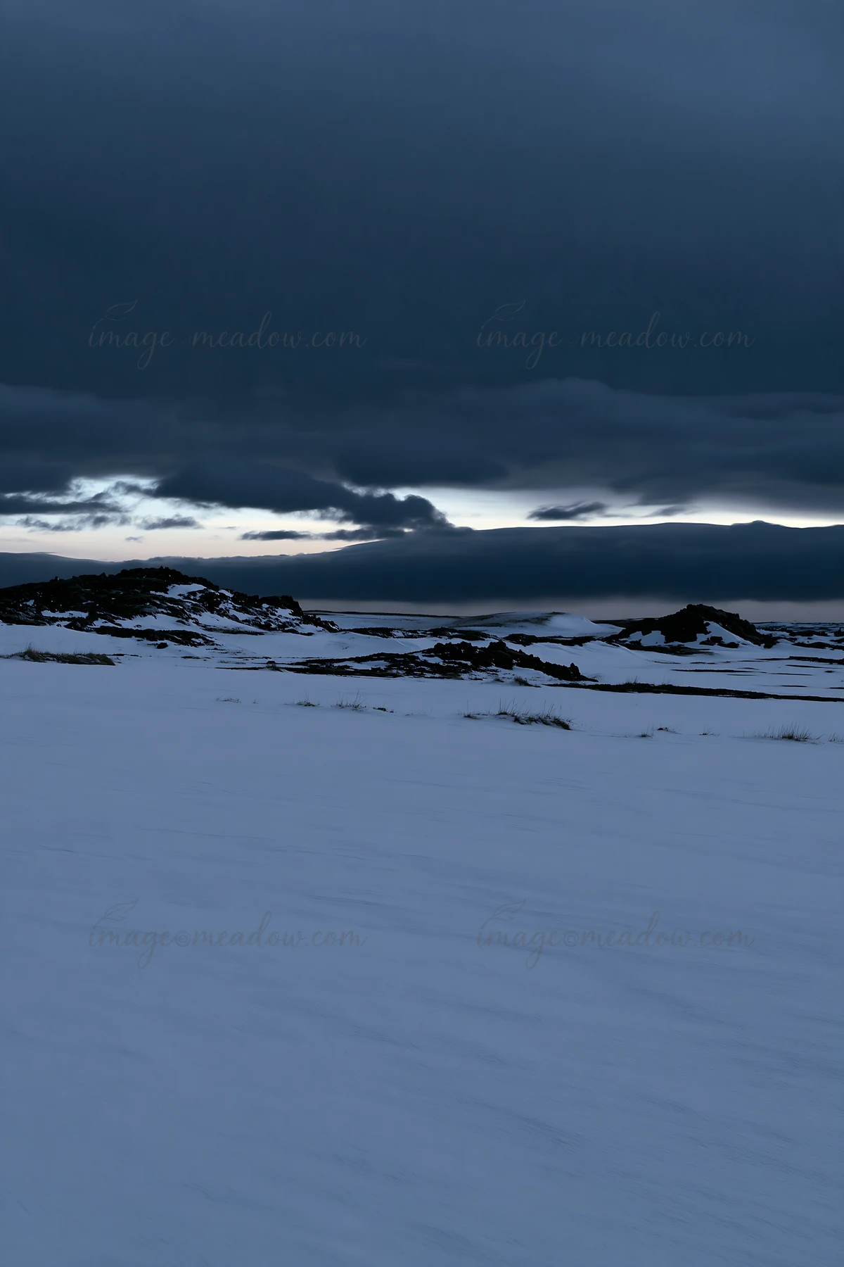 A snow-covered meadow stretches across a winter landscape. Dark clouds fill the sky, creating a dramatic scene above the white terrain. The location shows littl