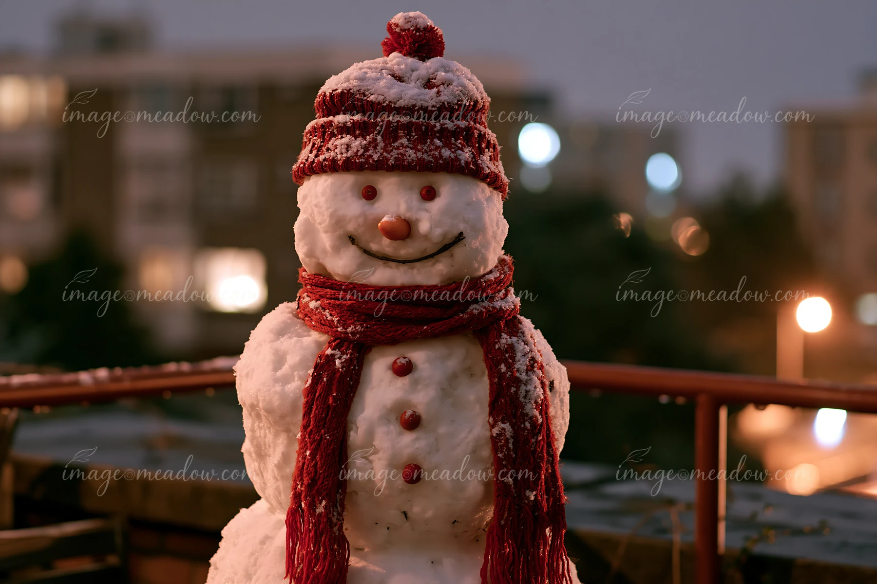 Snowman in a red hat and scarf stands on a snowy rooftop at night with blurred city lights behind it.