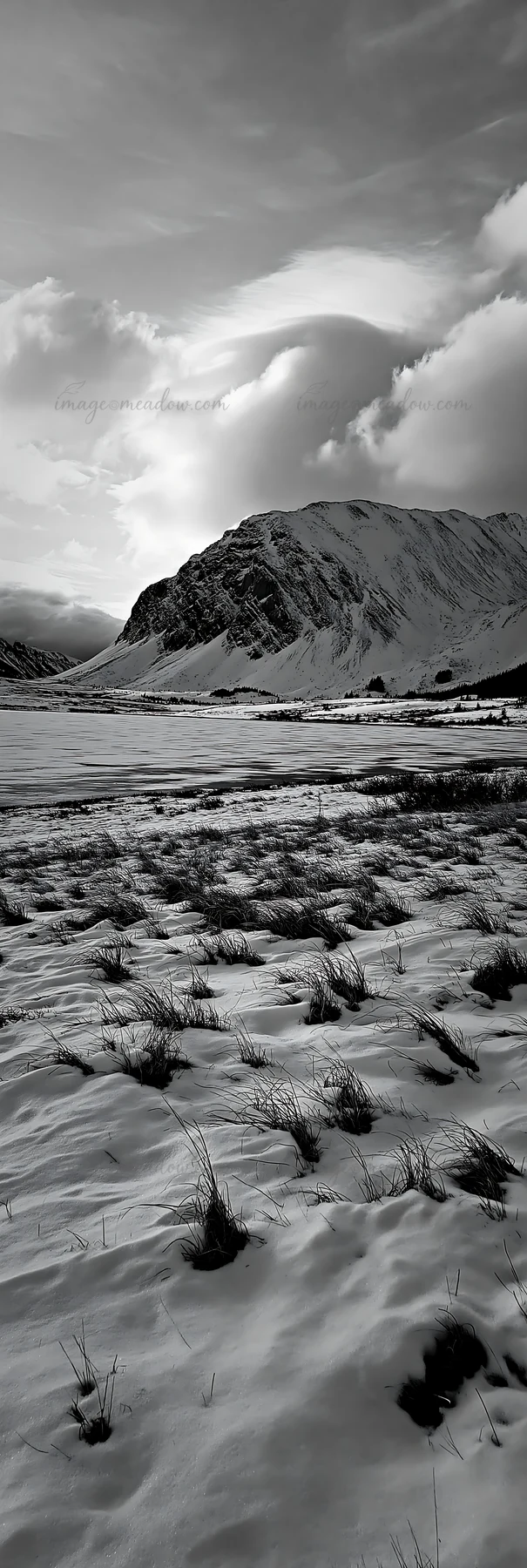 A snowy meadow borders a lake with mountains in the distance. Cloudy sky above adds depth to the scene. Visible grass peeks through the snow, creating contrast