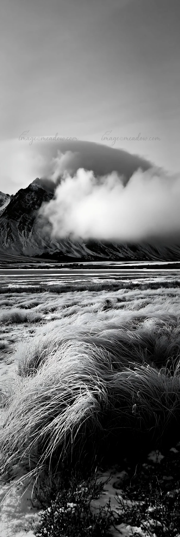Snow covers a meadow near a lake. Mountains rise in the background, partially hidden by clouds. The scene captures a quiet winter afternoon in a remote area.