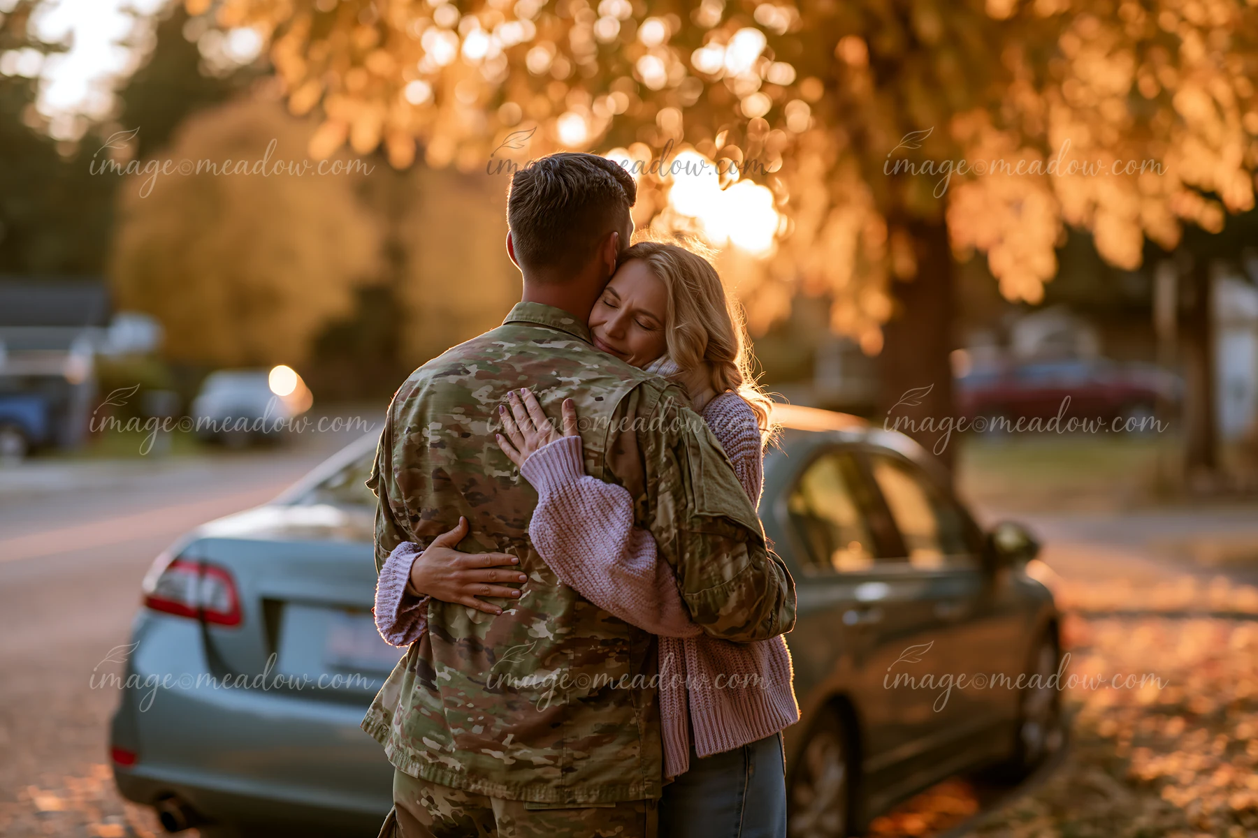 Soldier in camouflage embraces a woman beside parked cars on a residential street at sunset.