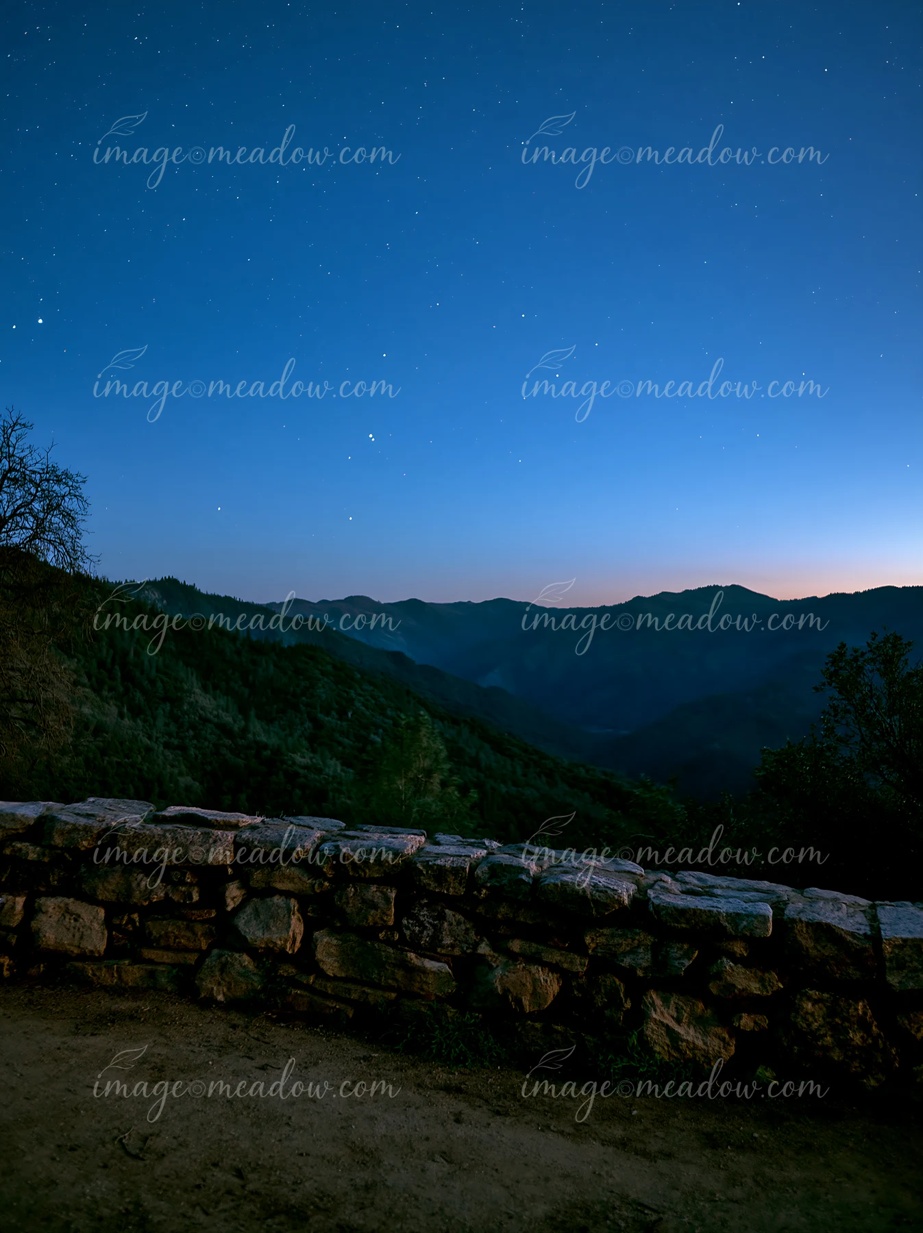 Stars fill the night sky above a mountain valley, with a stone wall in the foreground.