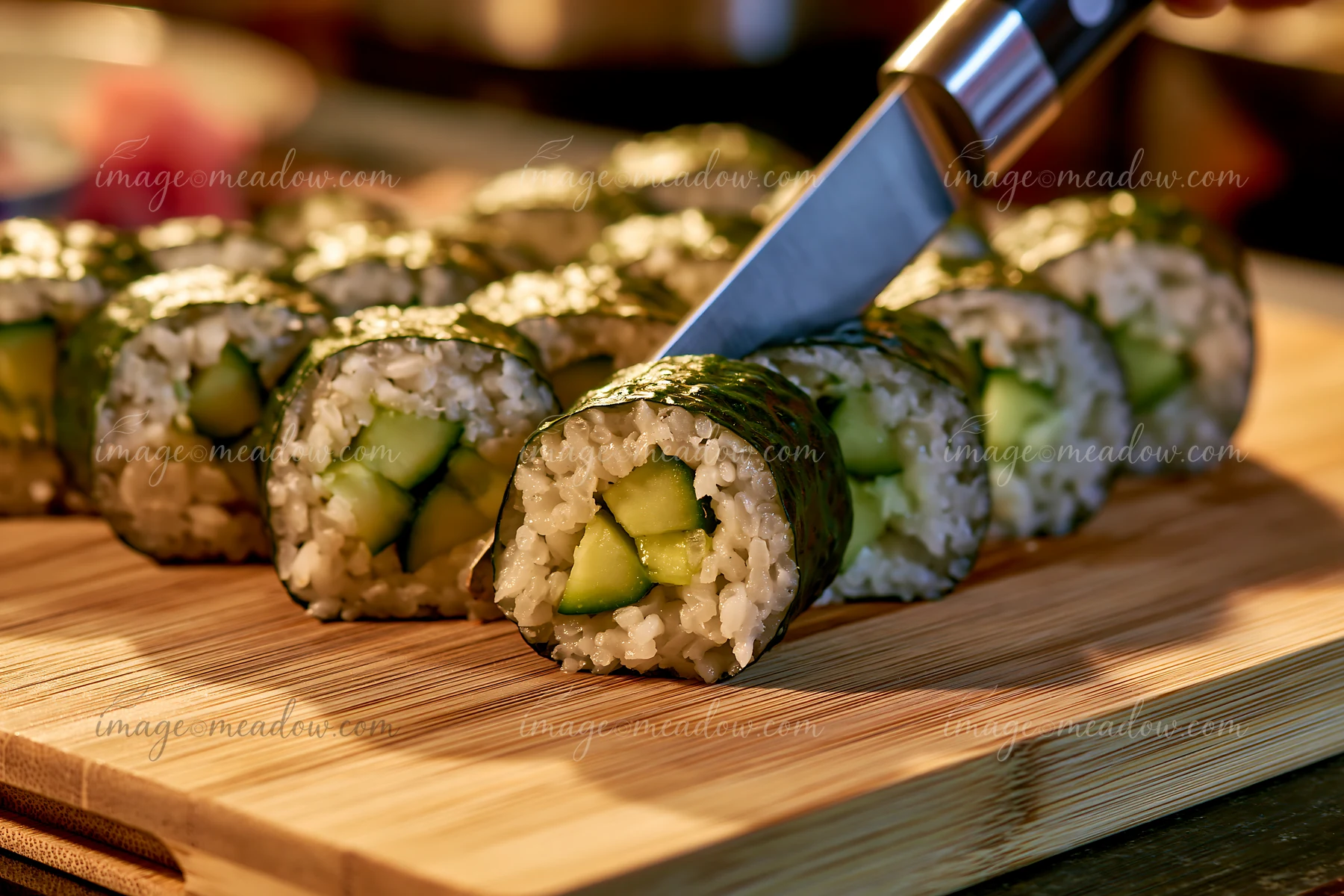 Sushi rolls being cut with knife on a wooden board in a kitchen setting.