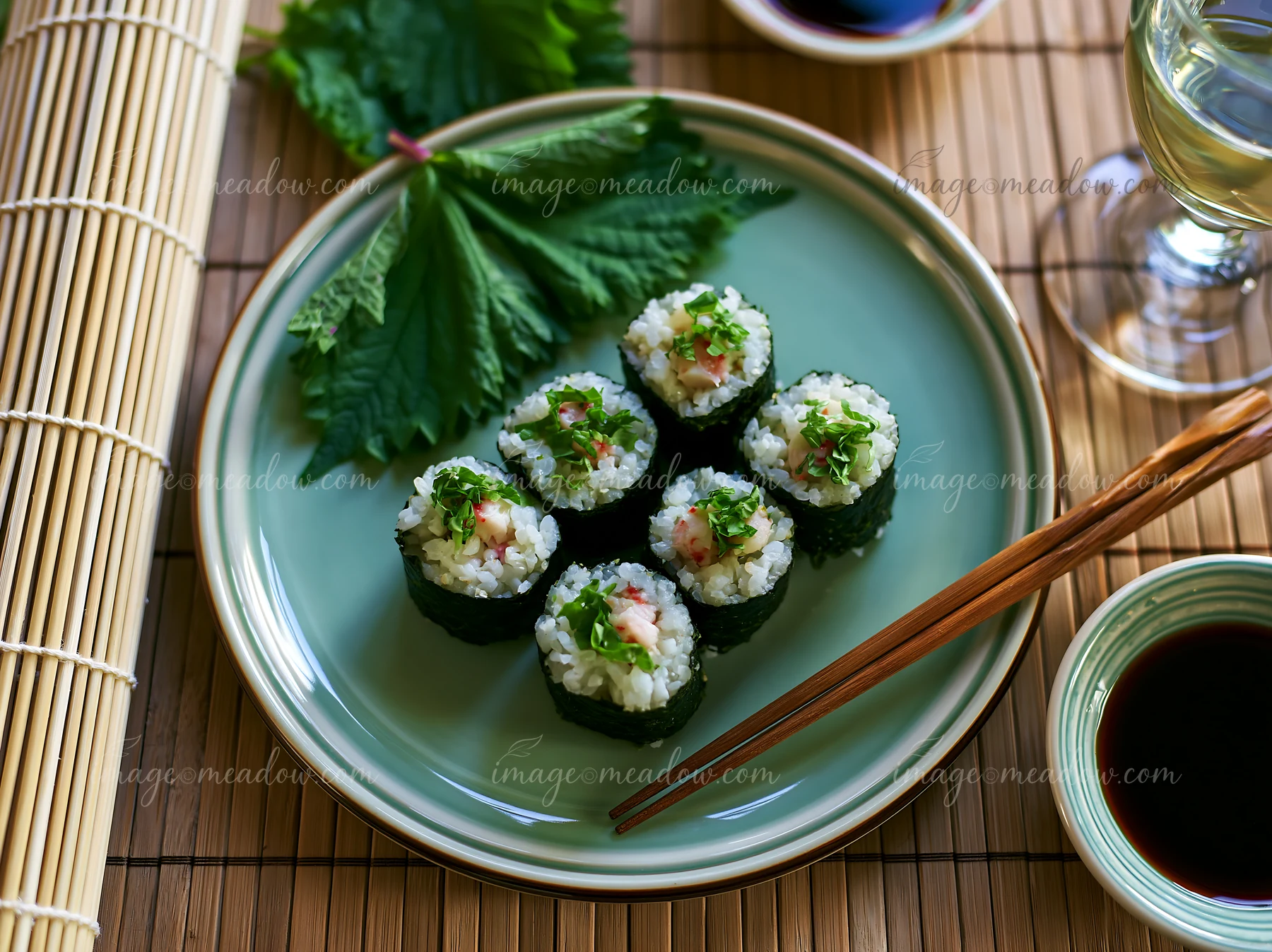 Sushi rolls with shiso leaves sit on a green plate beside soy sauce and a glass of sake.