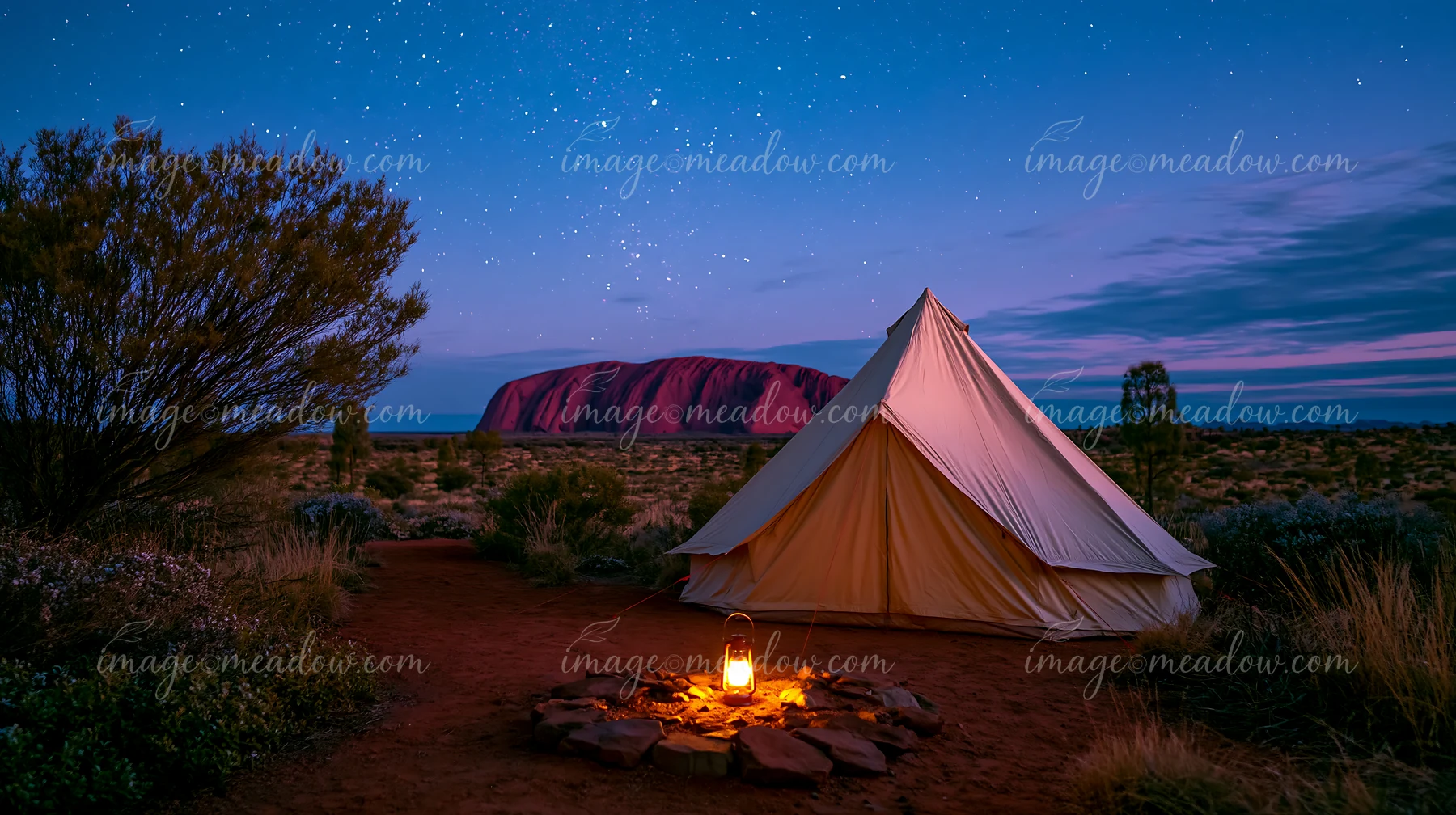 Tent and glowing lantern in an Australian desert camp with Uluru beneath a star-filled night sky.