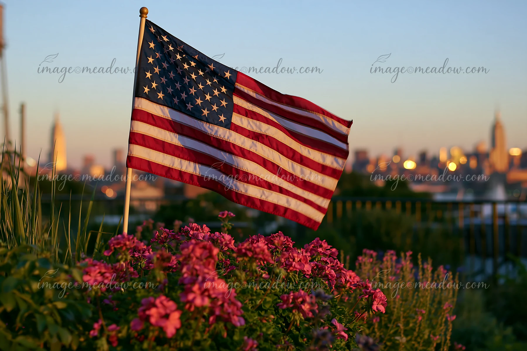 The American flag flying in strong wind stands out against a backdrop of city buildings with flowers in the foreground.