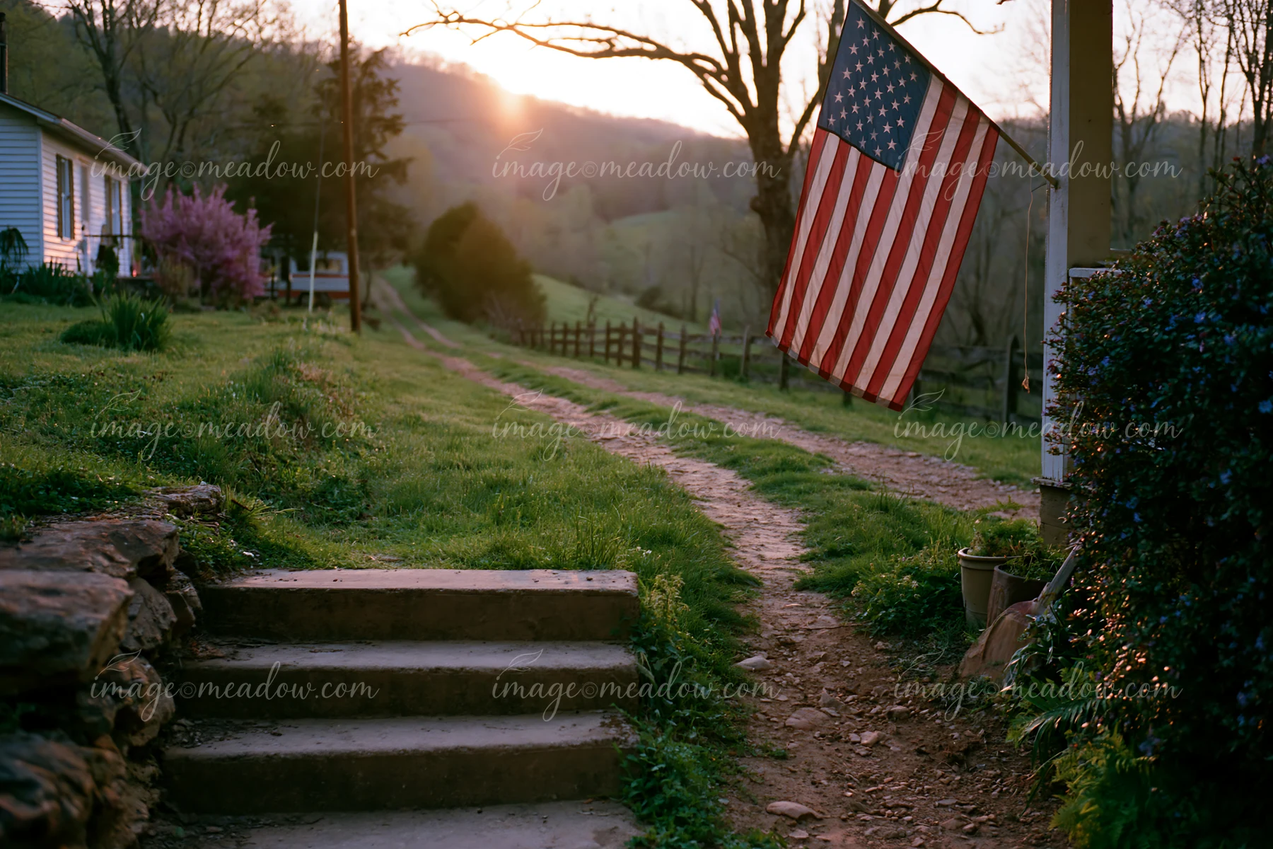 The American flag on rural road hangs from a porch as the sun sets over the hills.