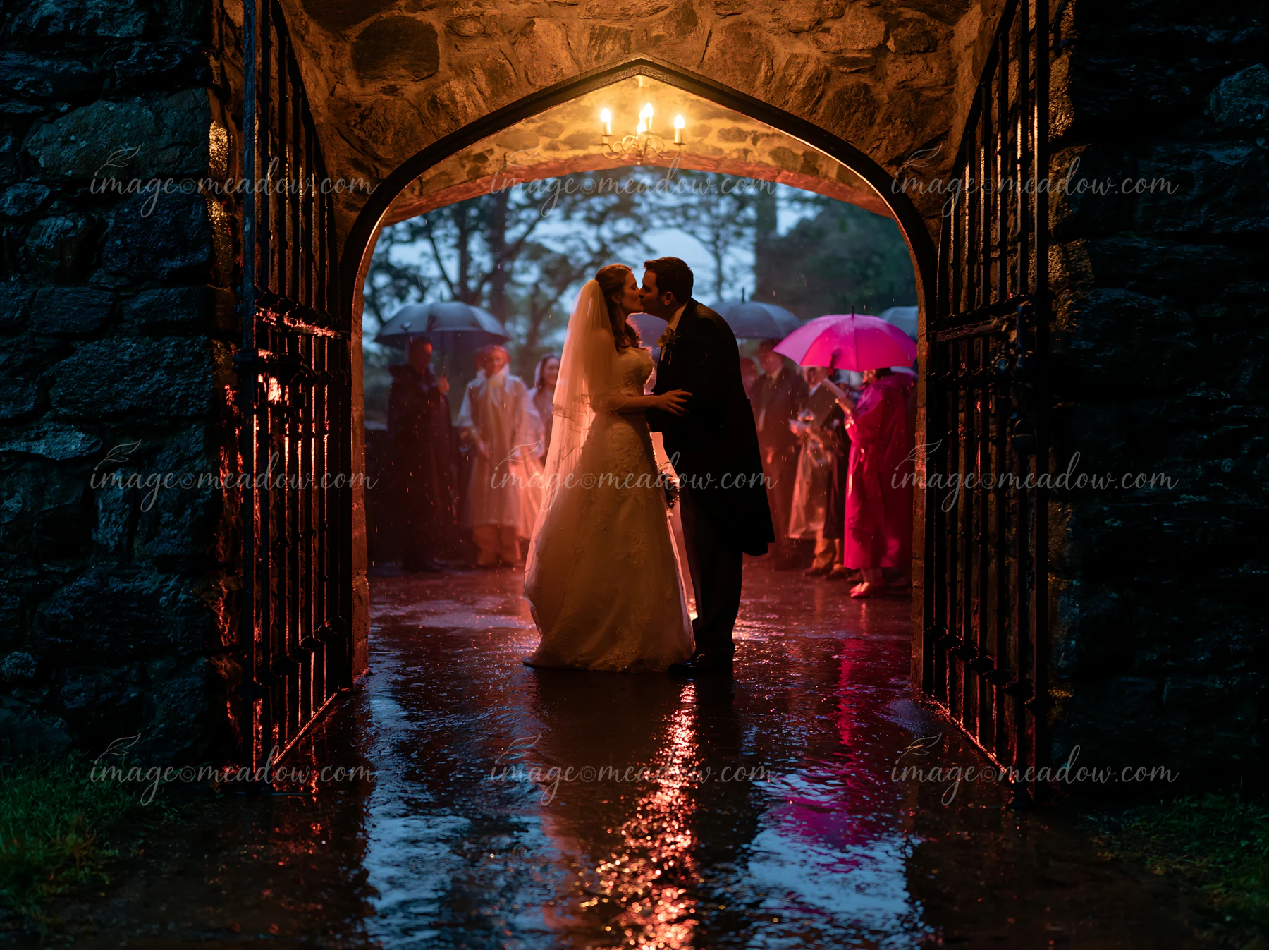 The bride and groom are kissing under an archway while guests hold umbrellas in the rain.