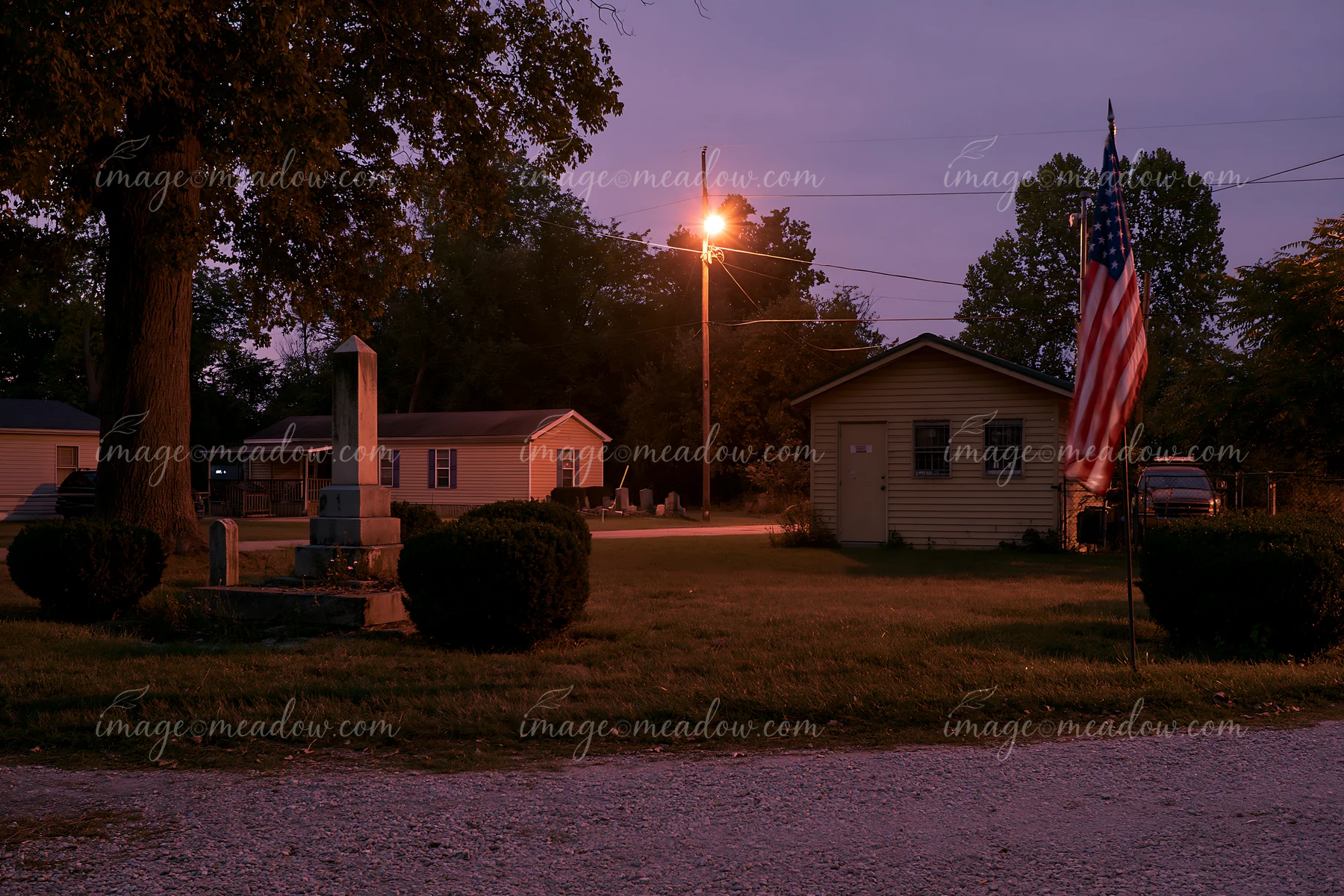 The cemetery monument and US flag appear in low light as evening falls, with a streetlight illuminating the area.