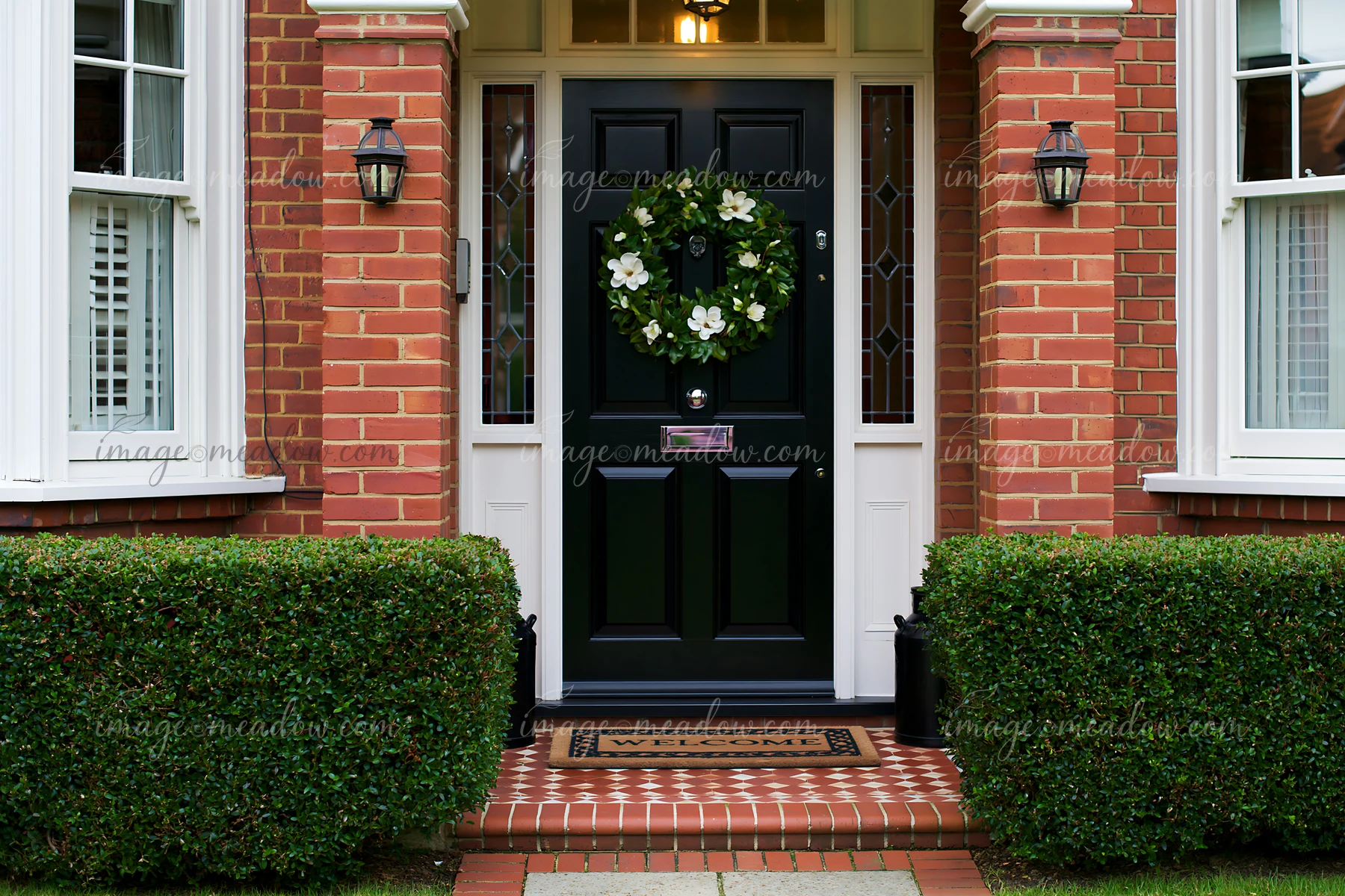 The front door with wreath and welcome doormat features a black door framed by brick walls and greenery.