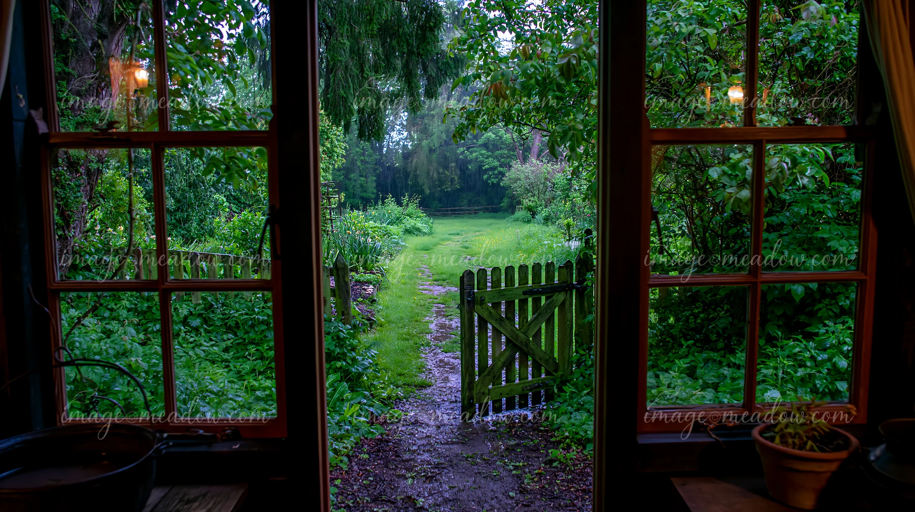 The scene shows a rainy day view from a timber cottage, with a path leading to a gate surrounded by lush greenery.