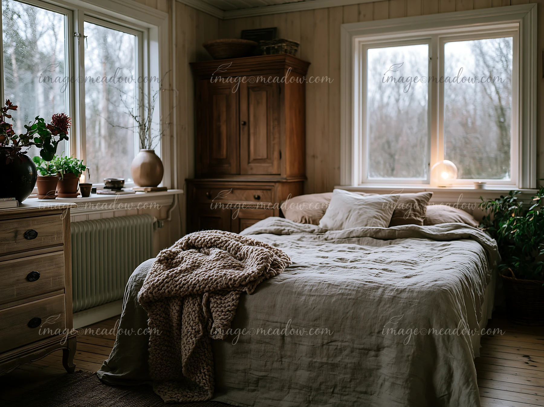 This rustic bedroom features a bed with linen bedding, wooden furniture, and large windows allowing natural light to fill the space.