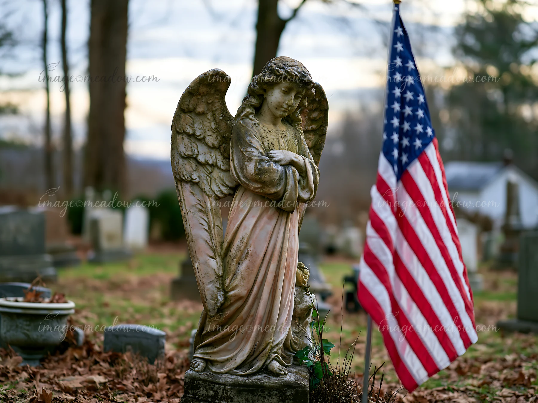 This solemn cemetery scene with American flag and stars shows an angel statue standing among gravestones, surrounded by fallen leaves during a cloudy day.