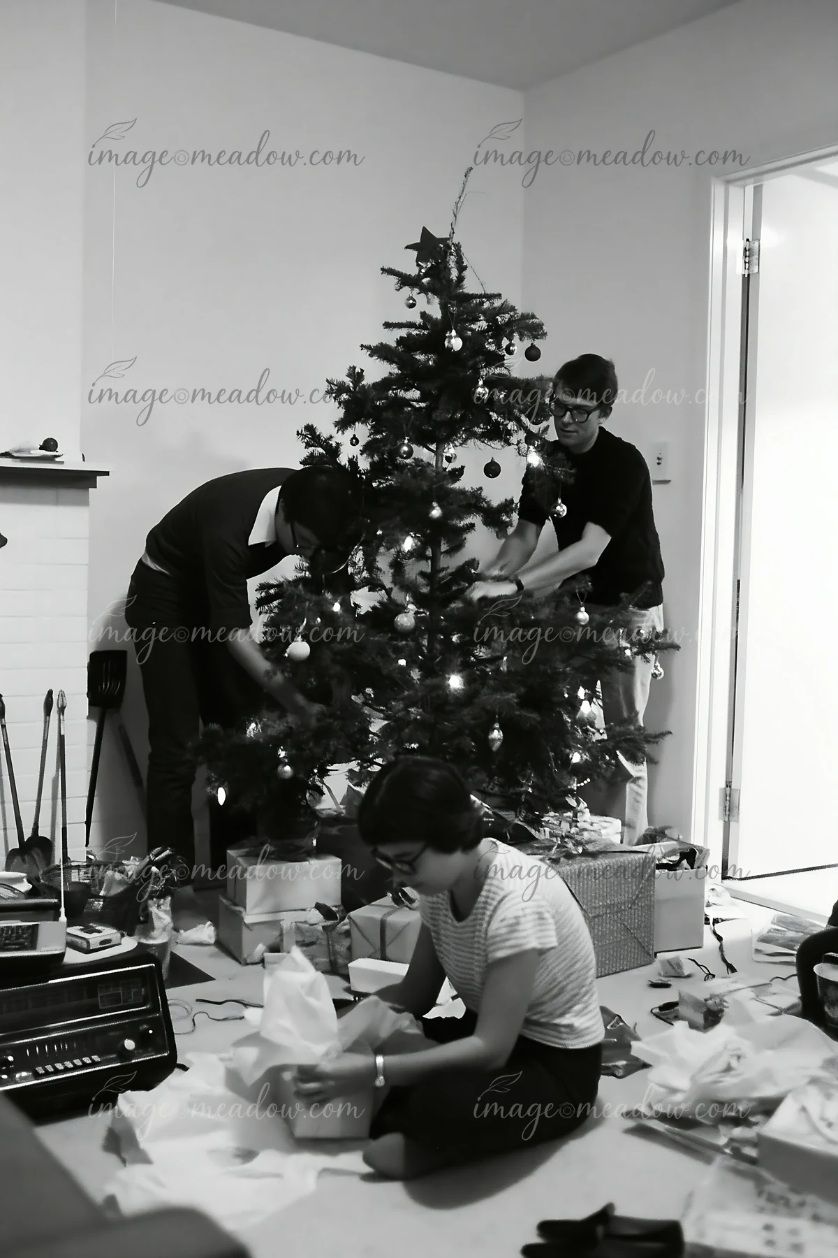 Three adults arrange wrapped gifts beneath a decorated Christmas tree in a home.