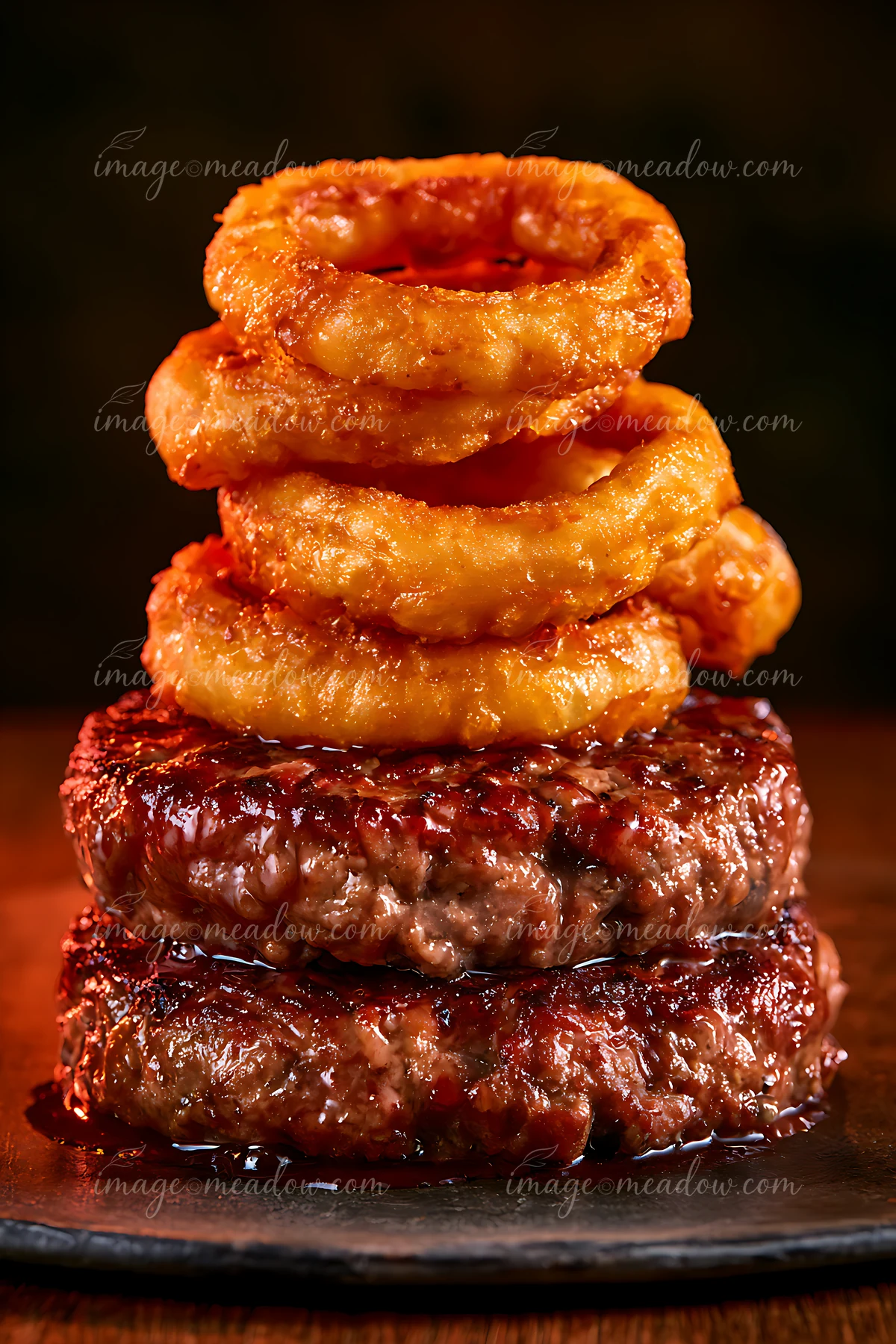 Three beef patties stacked on a dark plate with crispy onion rings on top.