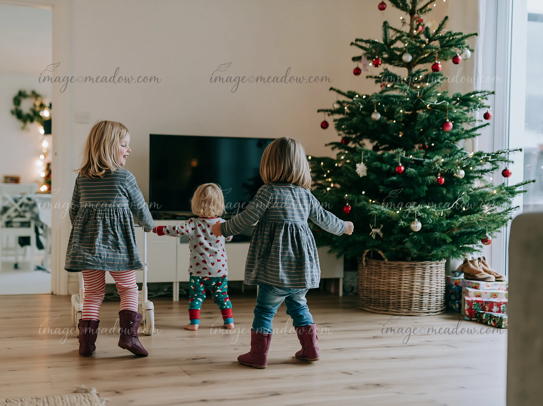 Three children dance joyfully around a decorated Christmas tree in a cozy living room.