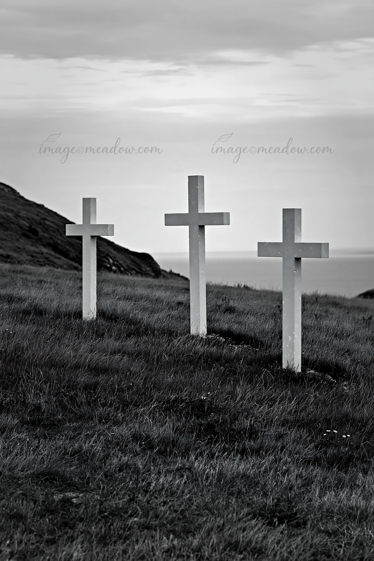 Three Christian crosses stand in a quiet meadow. The crosses are placed on grassy land, overlooking the sea. The scene has a simple and natural look, under a cl