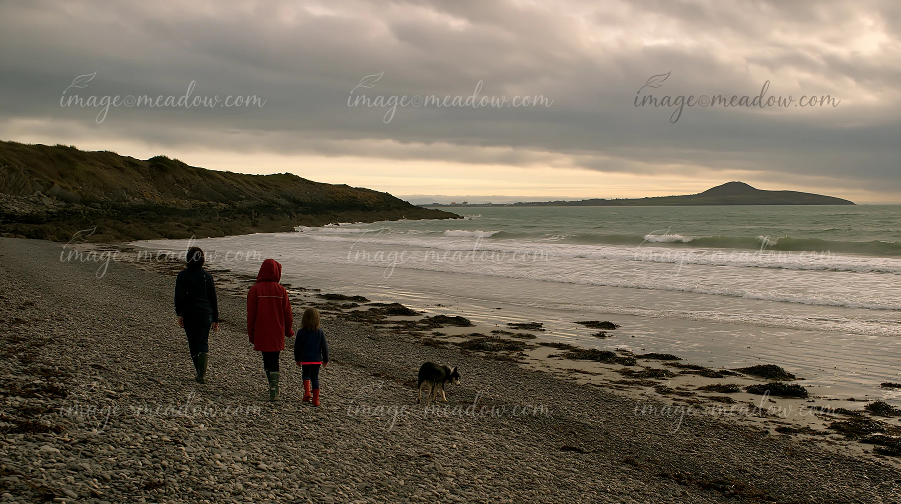 Three people and a dog walk along a rocky beach as waves wash ashore.