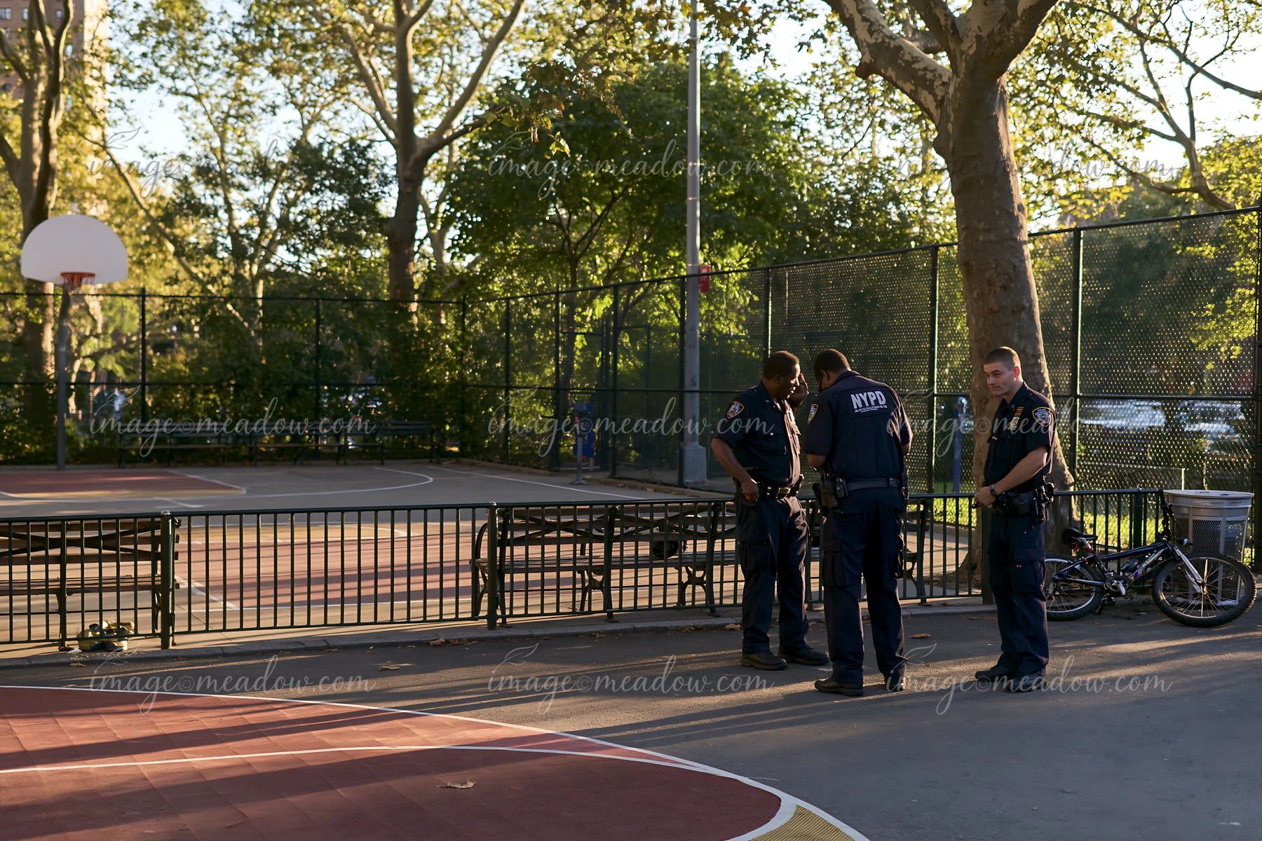 Three police officers are examining an area in a basketball court located in a public park during daylight hours.