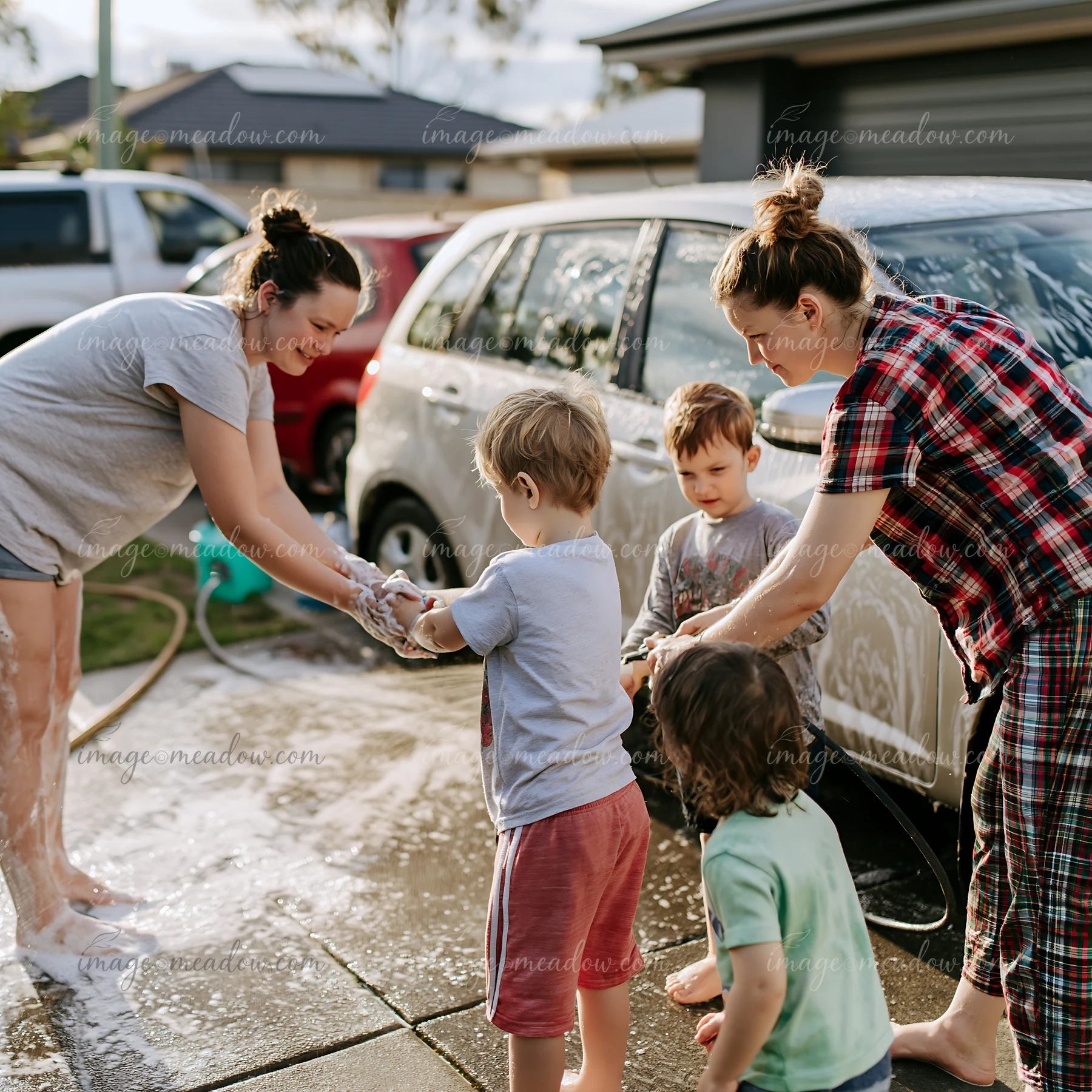 Two adults and three children wash a soapy white car together in a sunny residential driveway.