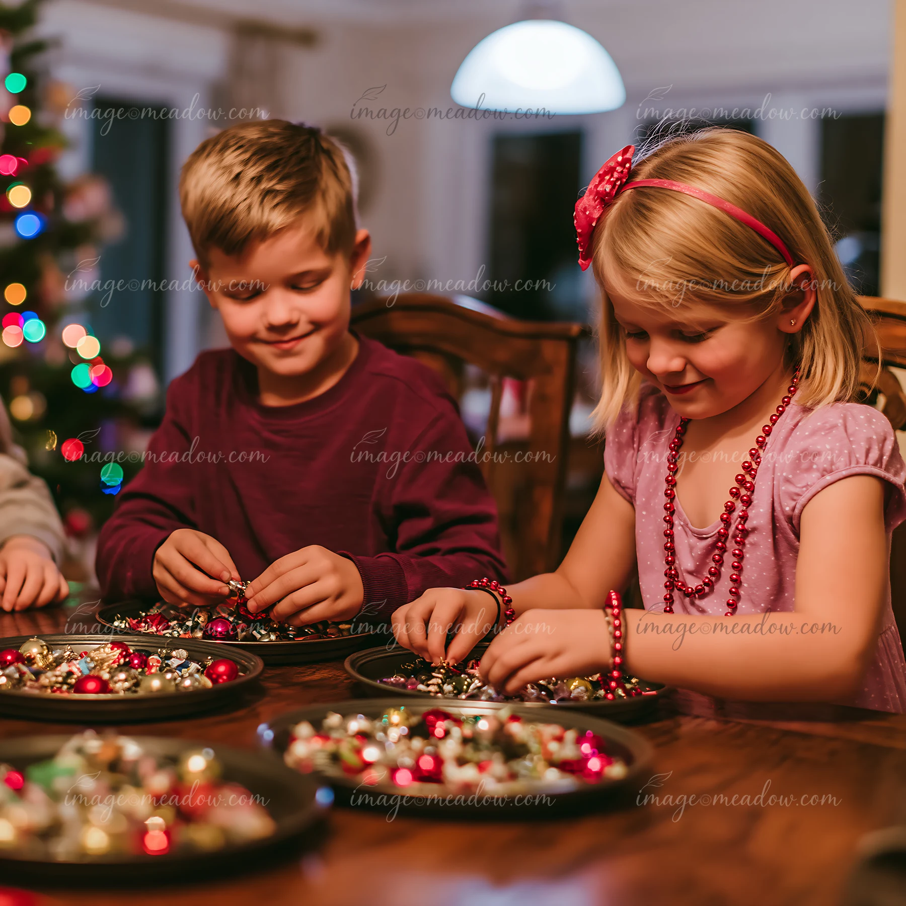 Two children are focused on sorting colorful Christmas decorations at a wooden table while holiday lights reflect in the background.