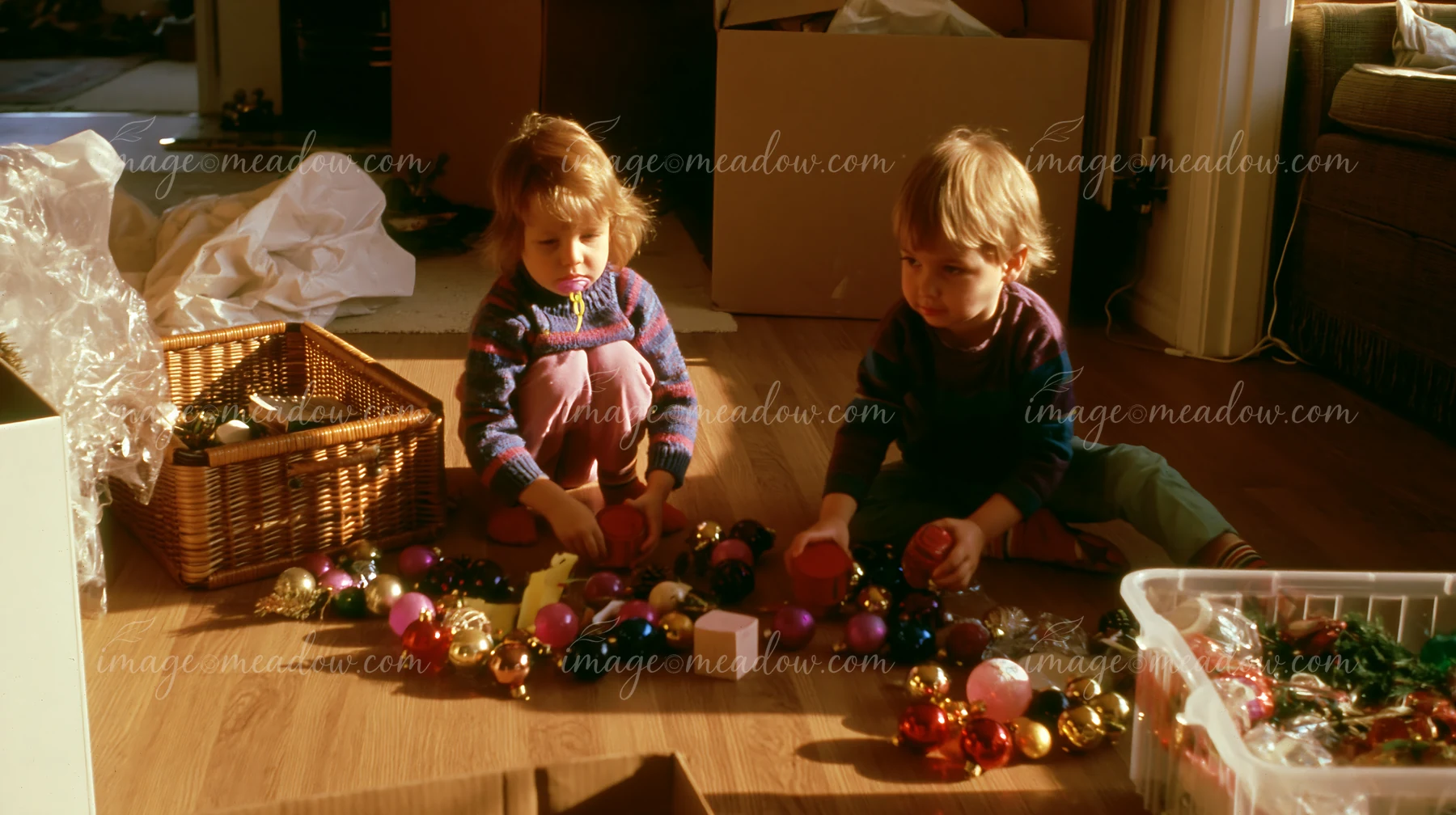 Two children are sorting Christmas decorations on the wooden floor of a living room.