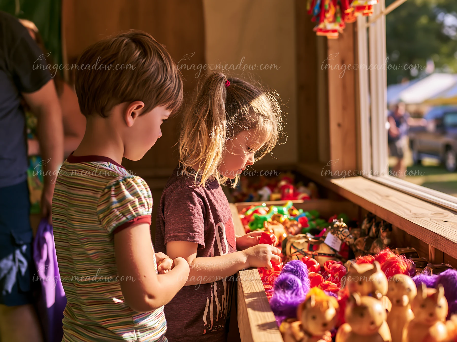 Two children choose colorful toys and dolls from a prize booth at a sunny outdoor carnival.