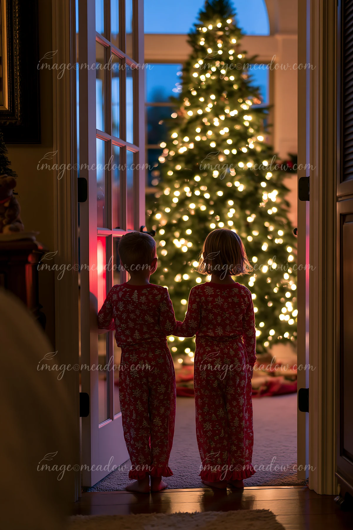 Two children in matching red pajamas stand in a doorway, looking at a brightly lit Christmas tree.