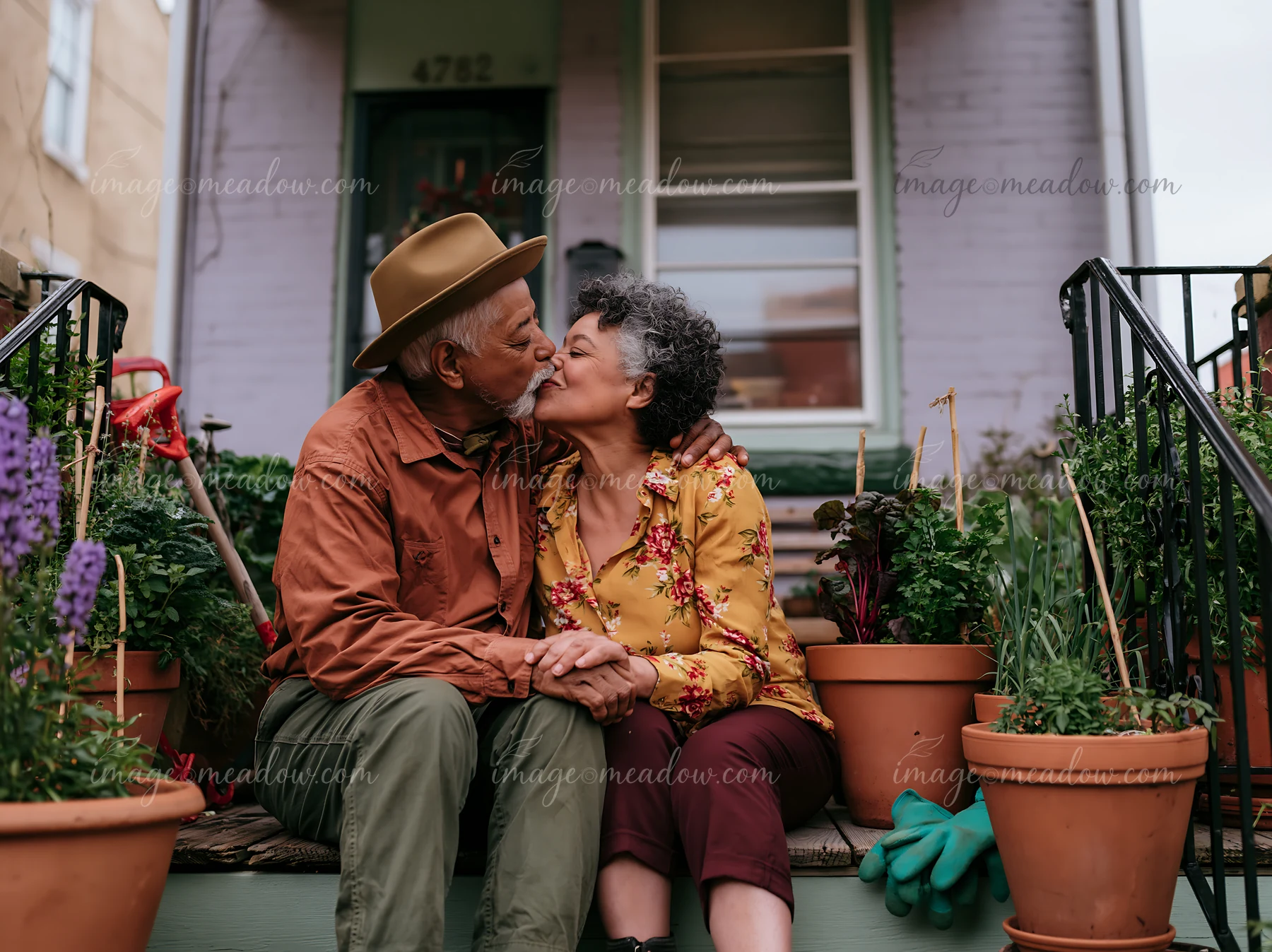 Two elderly partners share a kiss while sitting on their porch surrounded by potted plants.