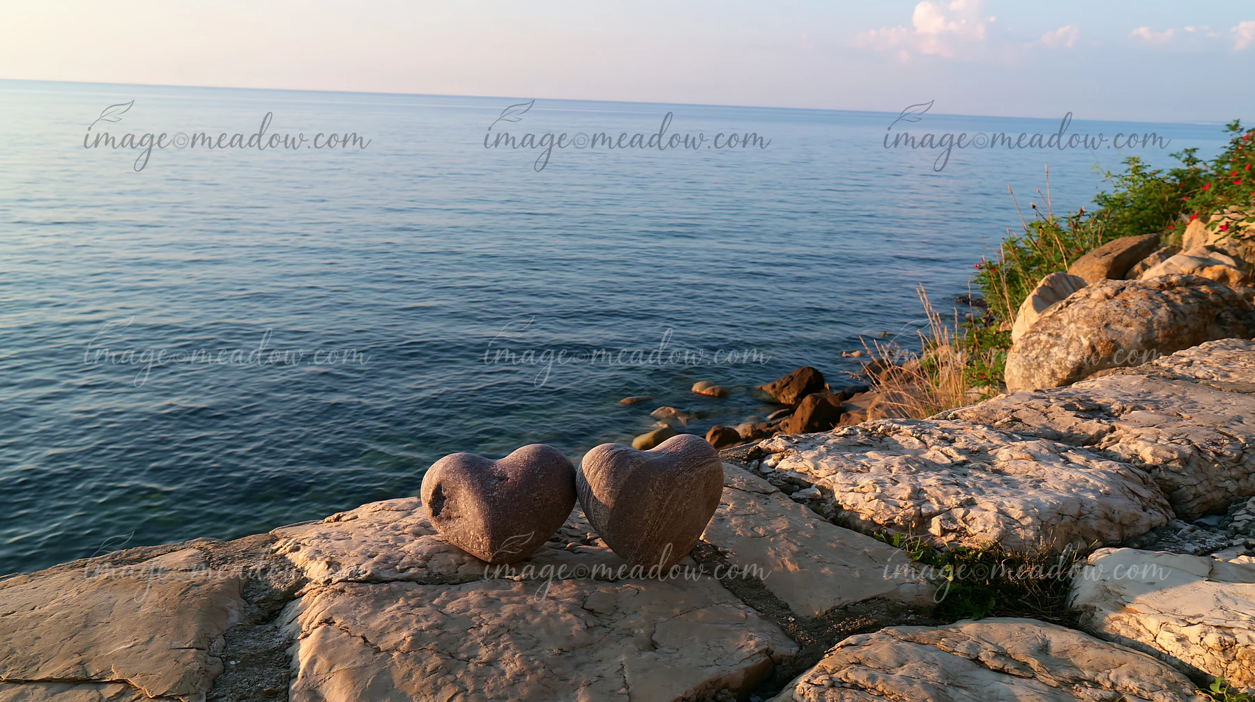 Two heart-shaped stones rest on sunlit coastal rocks beside calm blue ocean water.