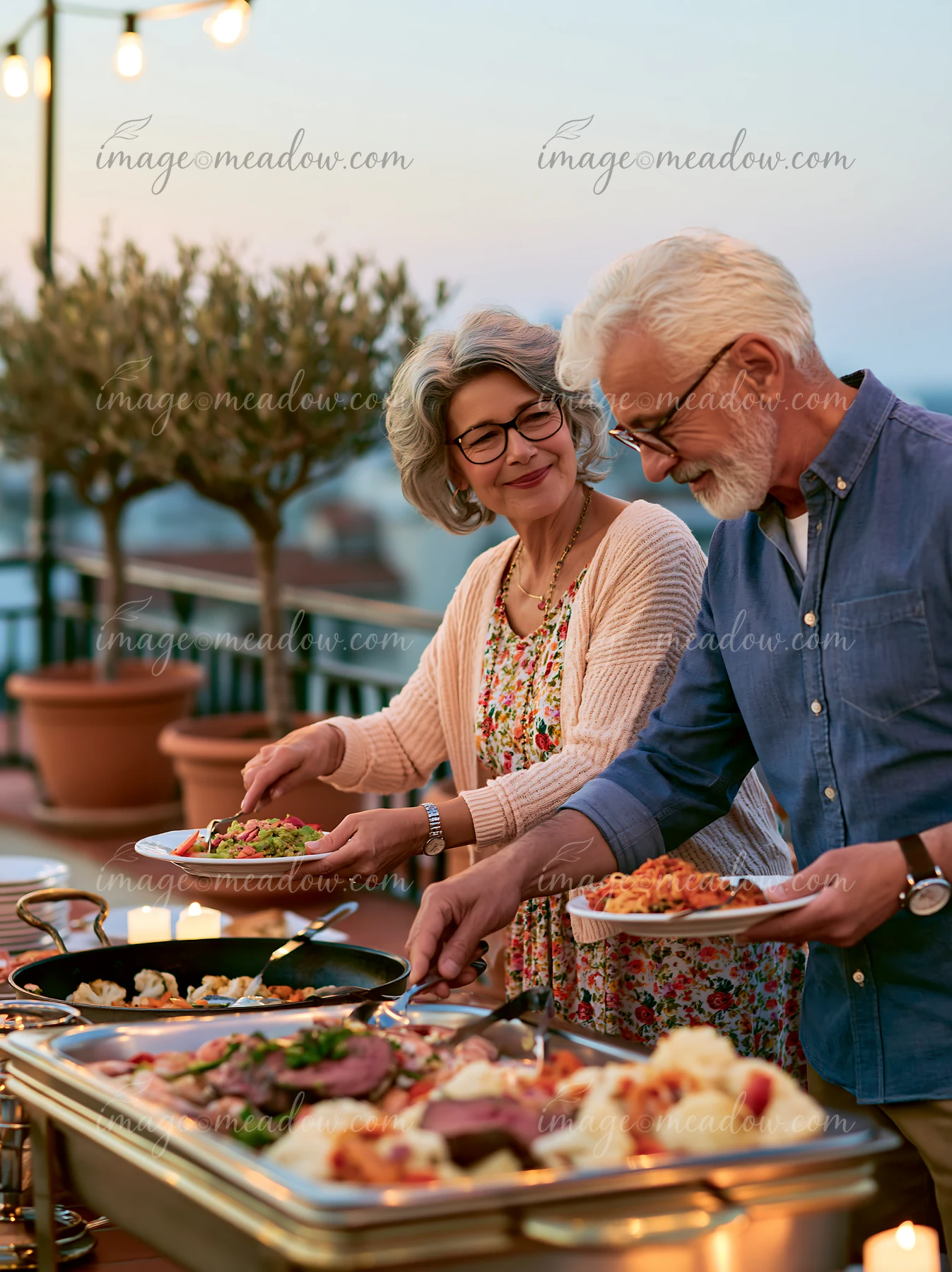 Two older adults serve themselves from a buffet during a rooftop dinner at sunset.