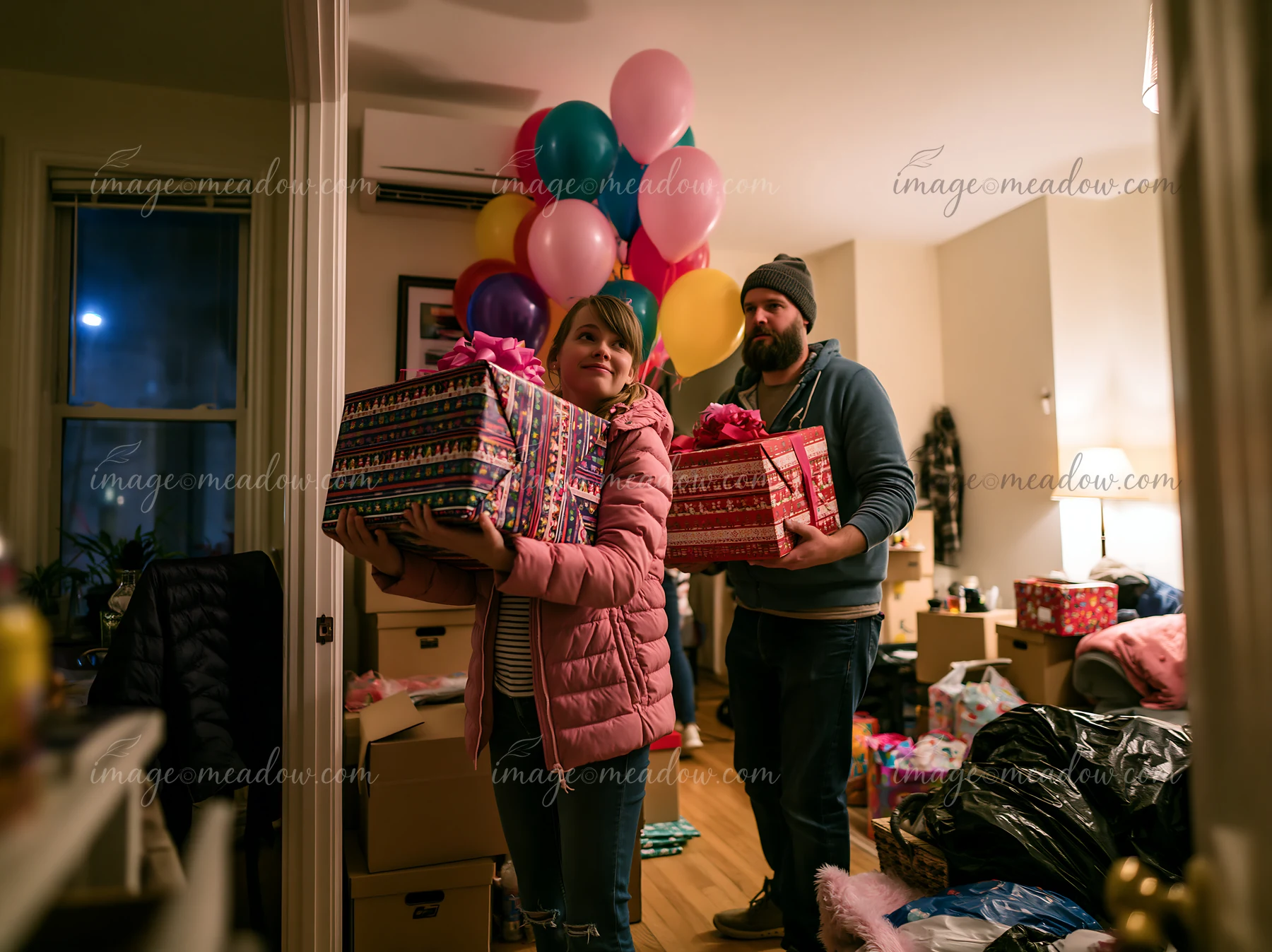 Two parents carry colorful decorated gift boxes through a warmly lit home filled with balloons.