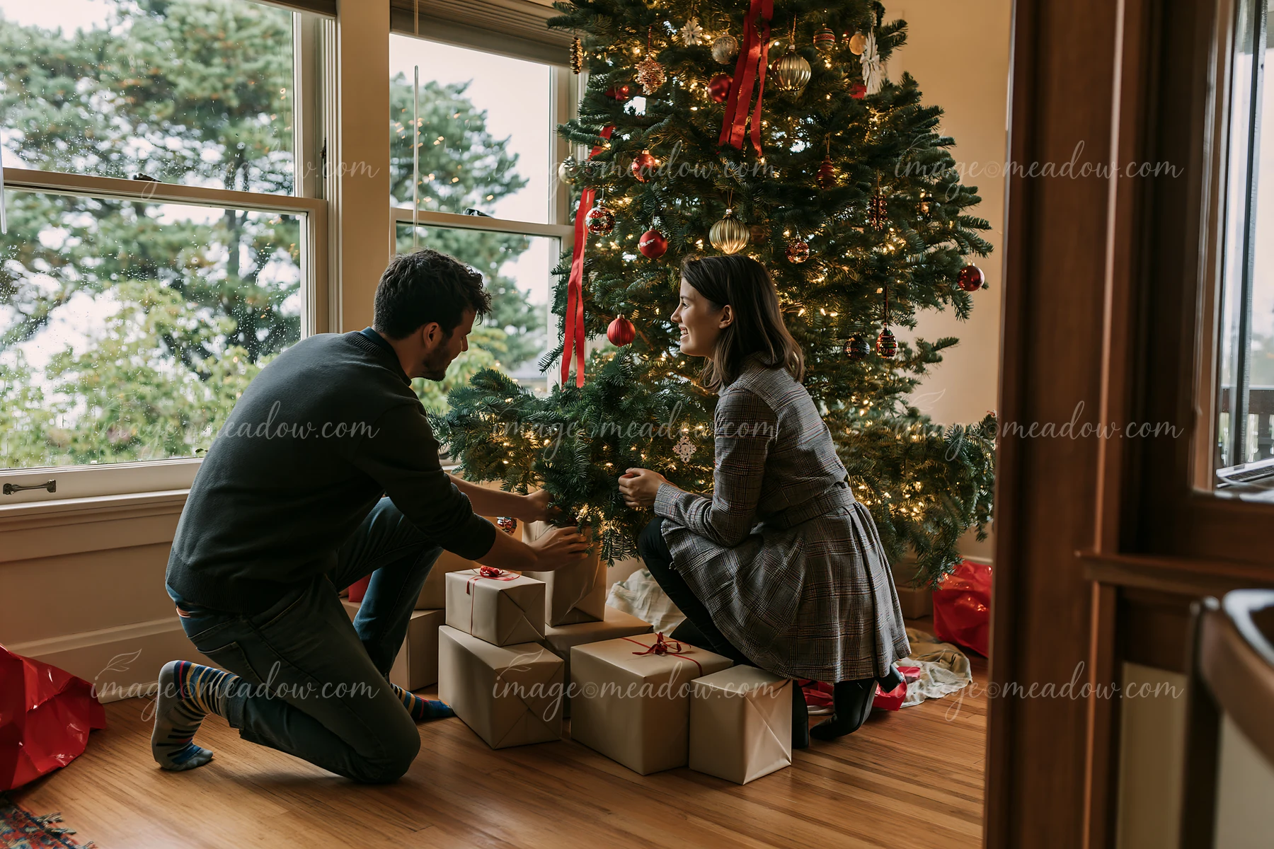 Two parents place wrapped gifts beneath a decorated Christmas tree in a warmly lit home.