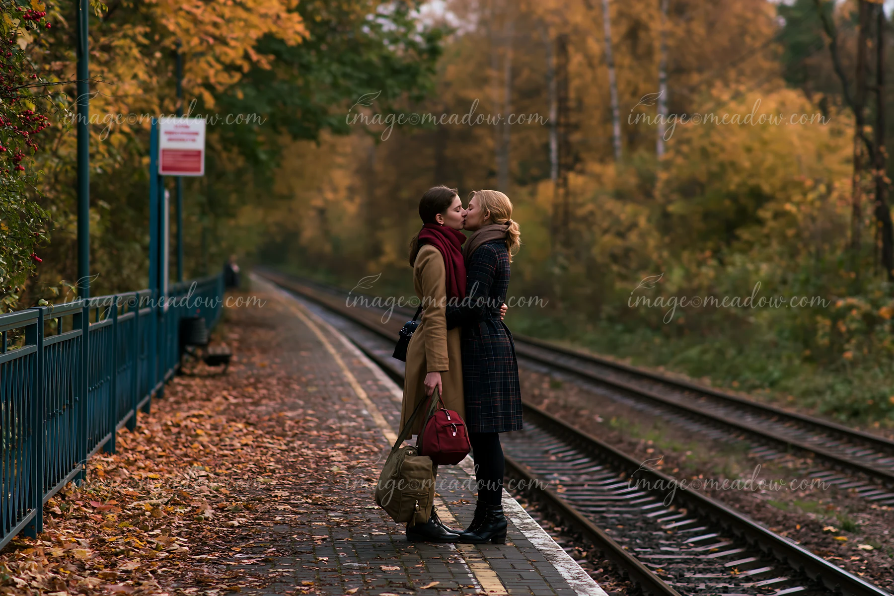 Two partners kiss beside railway tracks on a leaf-covered autumn train platform.