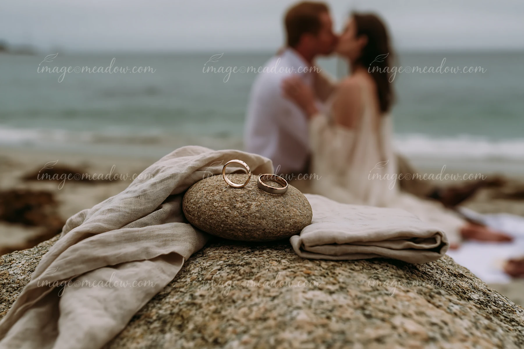 Two people kiss beside wedding rings resting on a rock at the beach.
