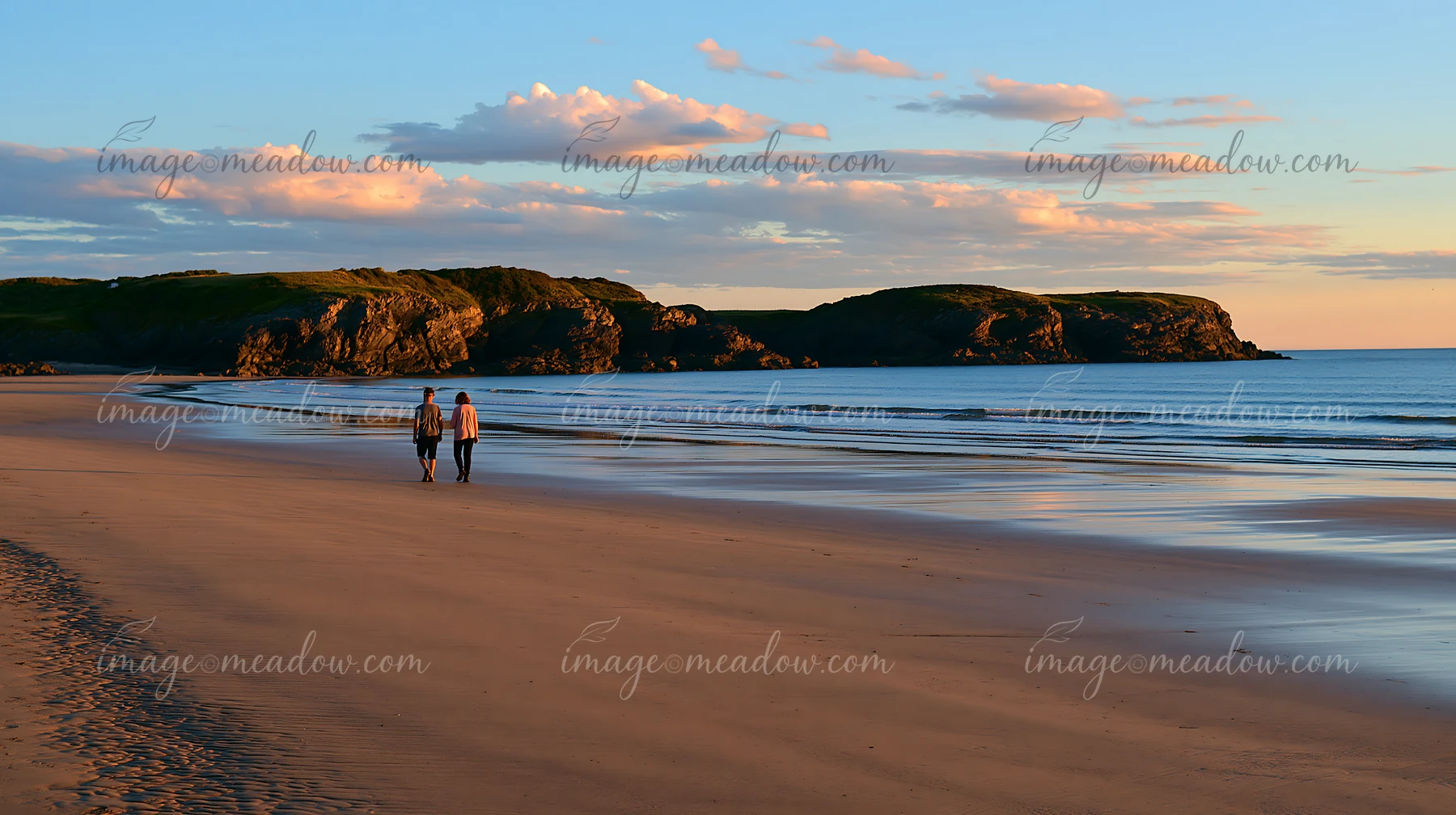 Two people walk along a sandy beach near a rocky peninsula as the sun sets in the background.