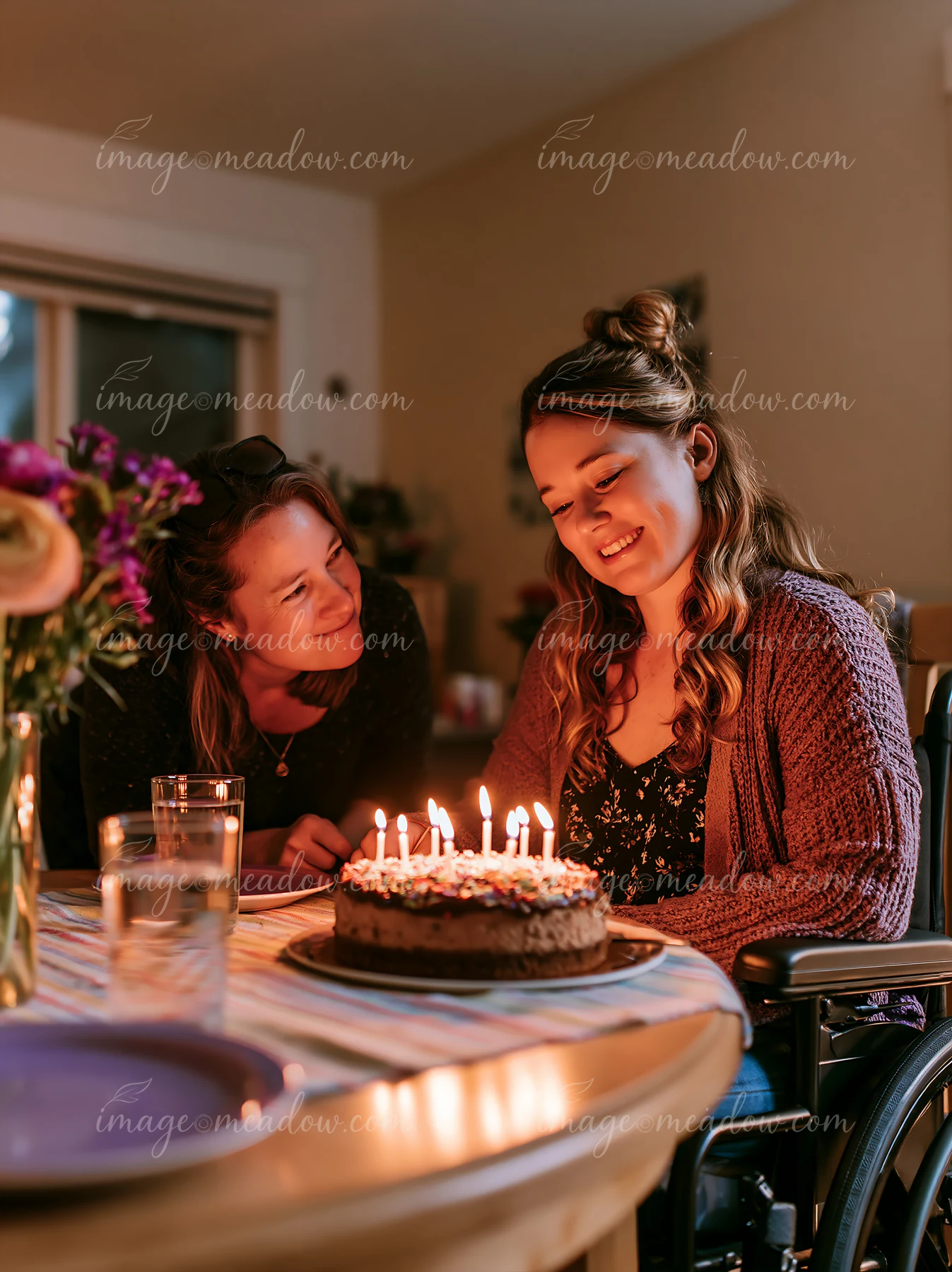 Two women light candles on a decorated birthday cake at a warmly lit dining table.