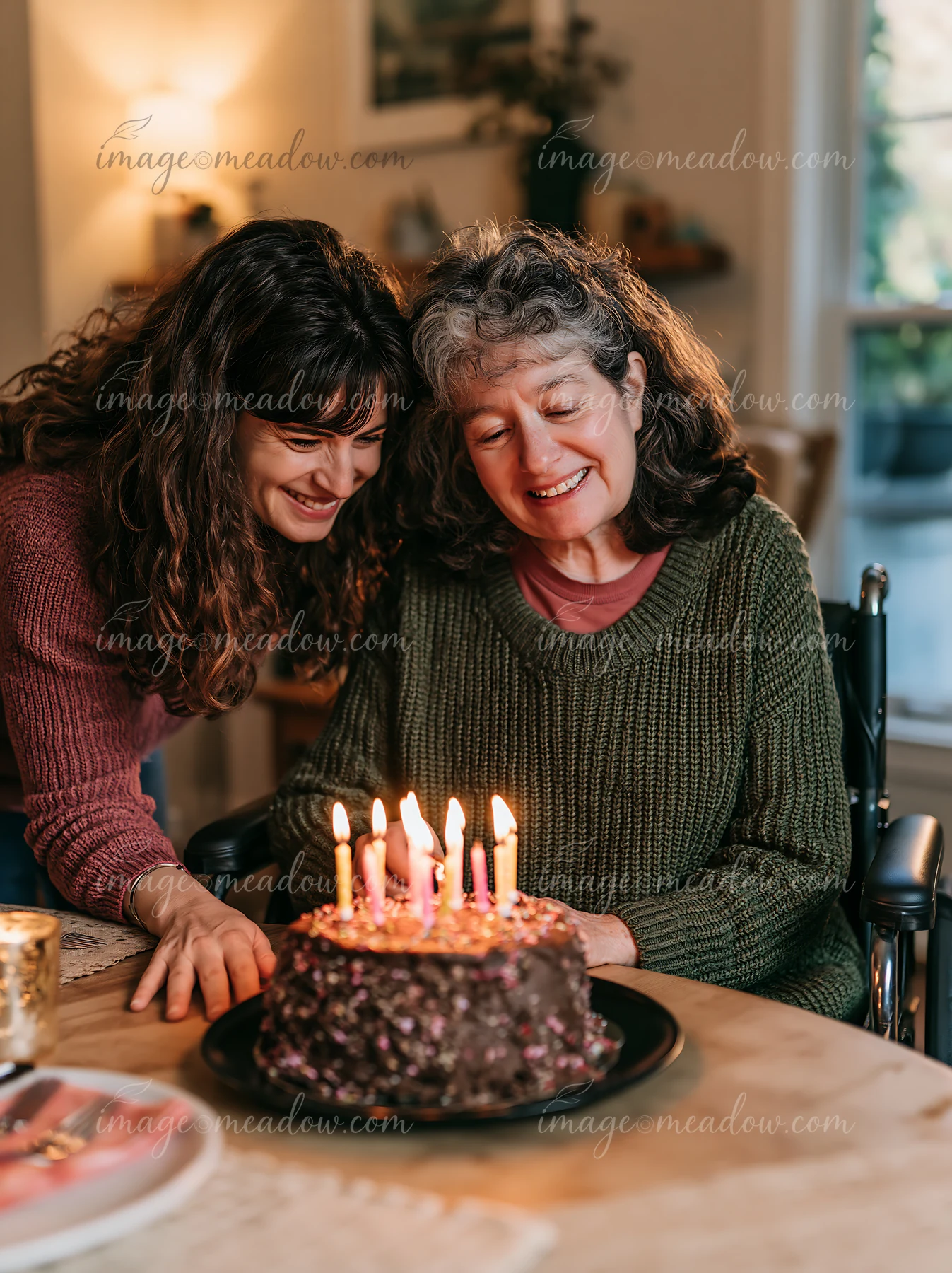 Two women smile beside a decorated birthday cake while candles are being lit at a dining table.