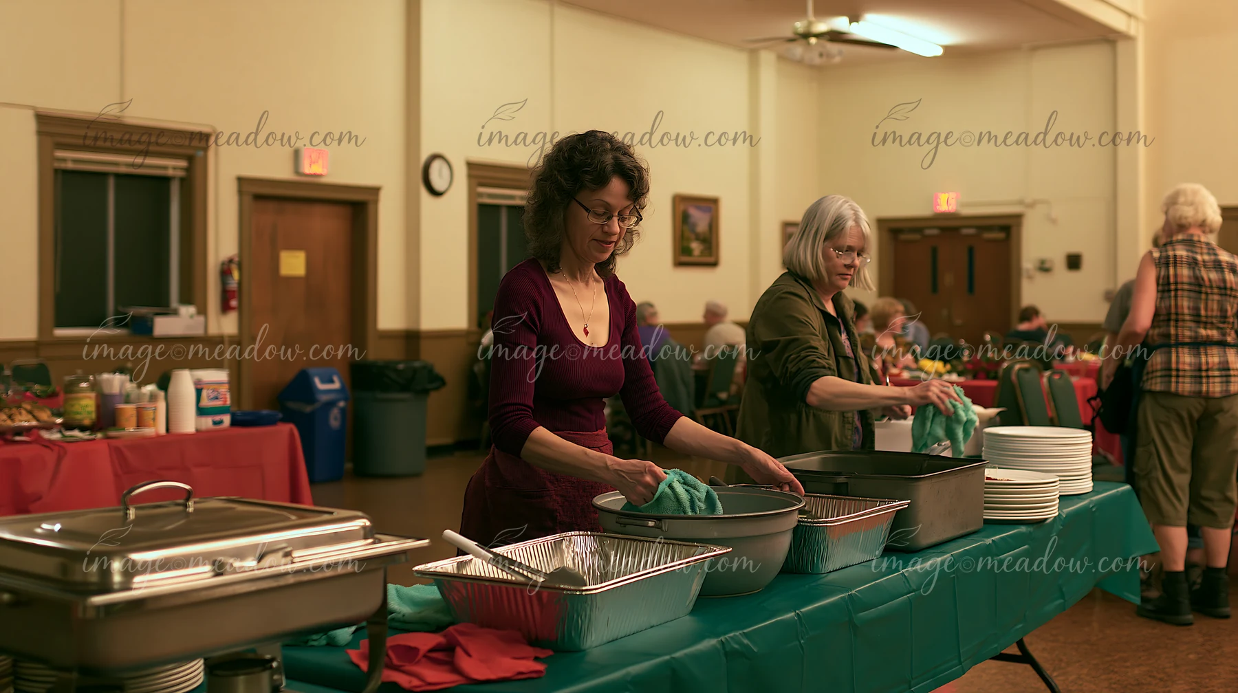 Two women work at a charity event buffet, arranging food and cleaning utensils in a community hall.