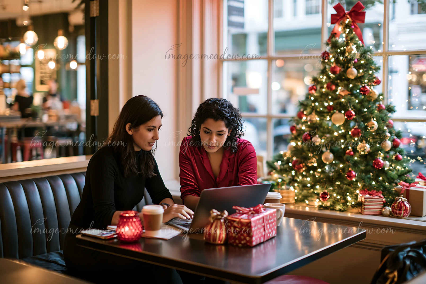 Two women work together at a coffee shop table beside a decorated Christmas tree and wrapped gifts.