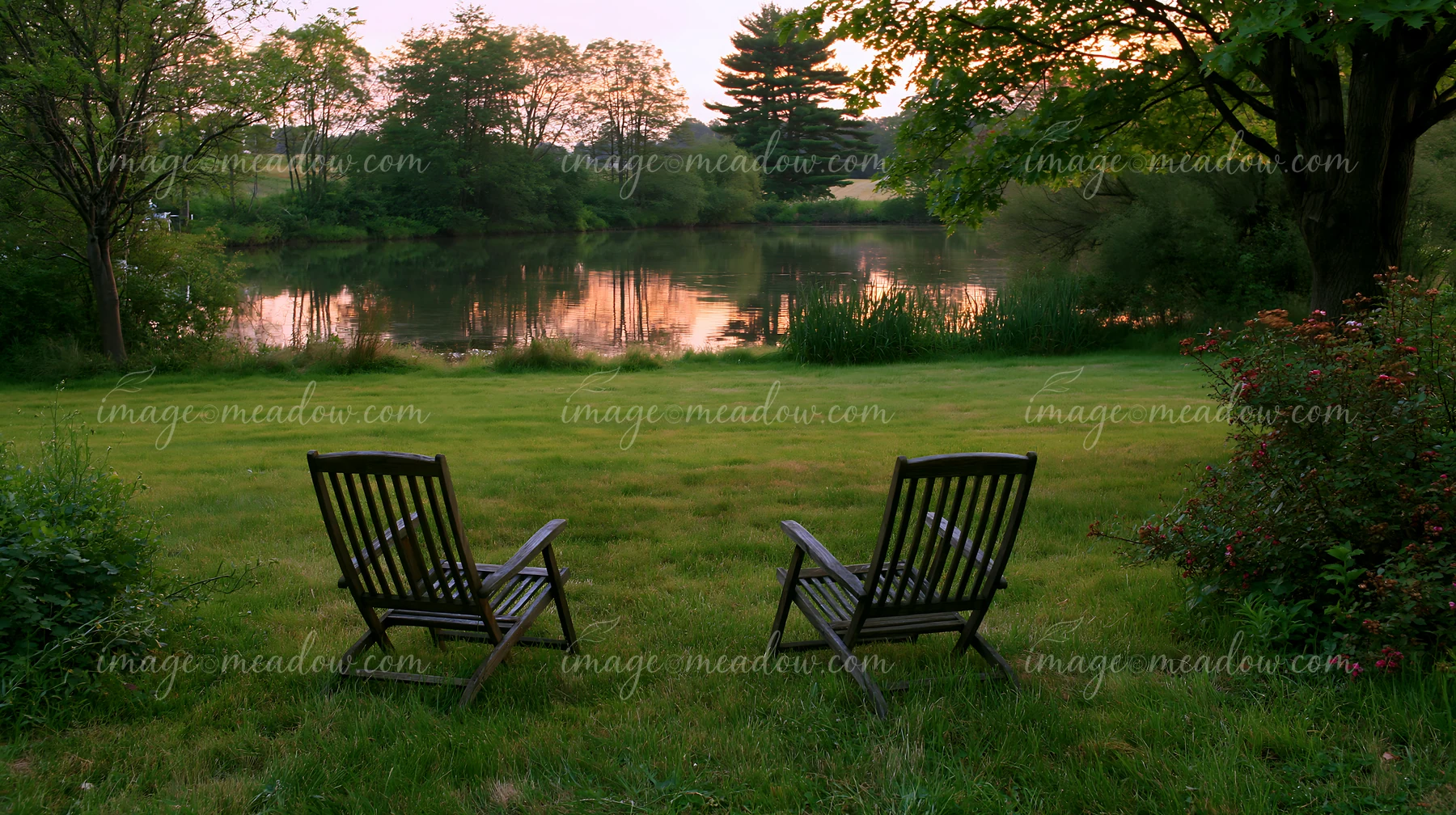 Two wooden chairs sit on a freshly mowed meadow beside a still lake, surrounded by trees.