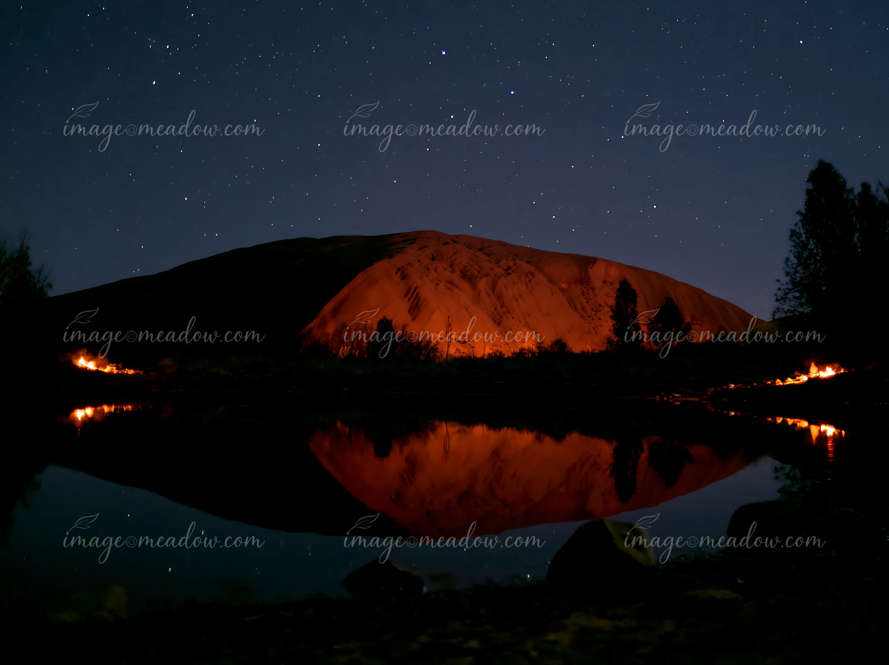 Uluru under a starry night sky showcases the large rock formation glowing under the starlight with reflections in a calm body of water nearby.