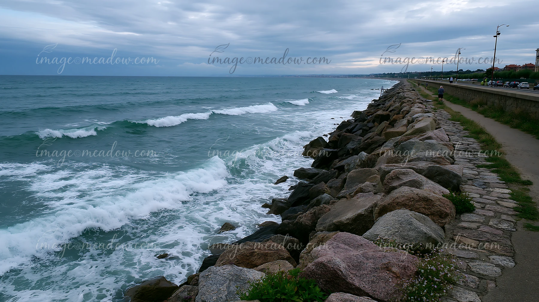 Waves break against a rocky sea wall beside a coastal road under a cloudy sky.