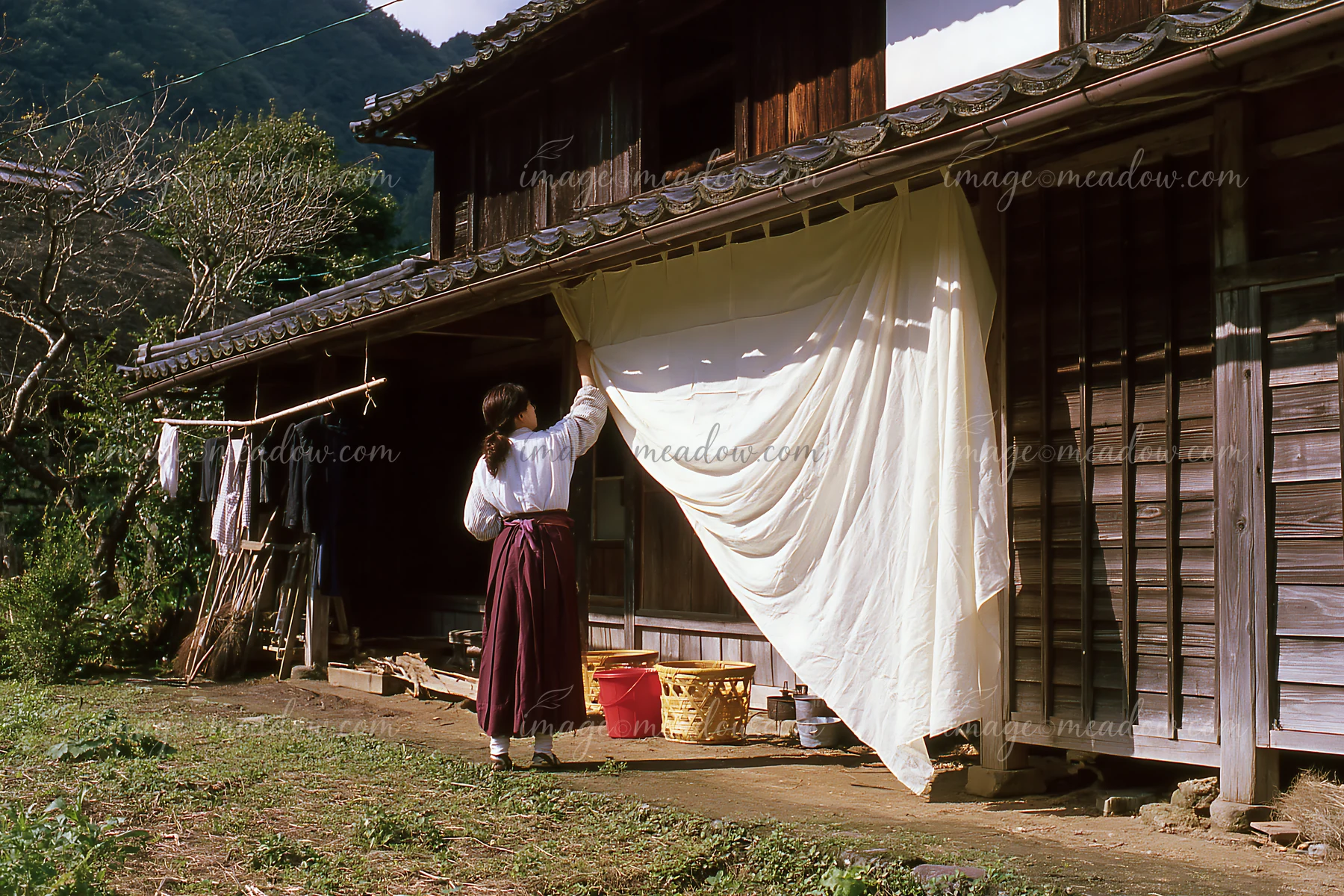 Woman in heritage attire hangs a large white fabric panel on a traditional wooden house.