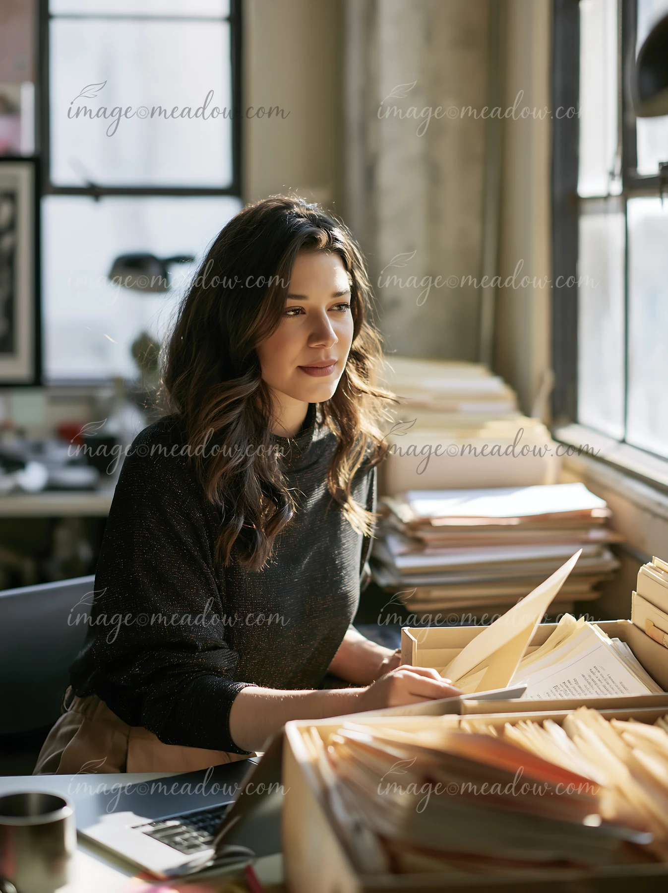 Woman sorts paper files at a desk beside a laptop in a sunlit office.