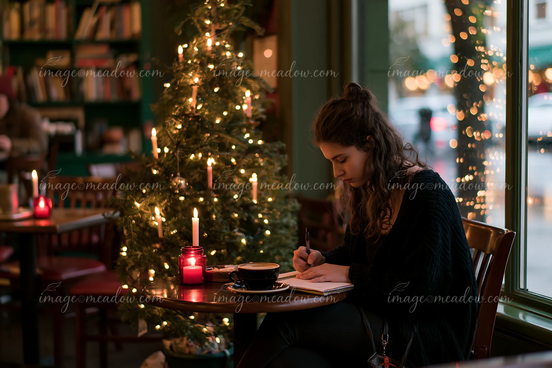 Woman writes in a notebook at a coffee shop table beside coffee, candlelight, and a decorated Christmas tree.